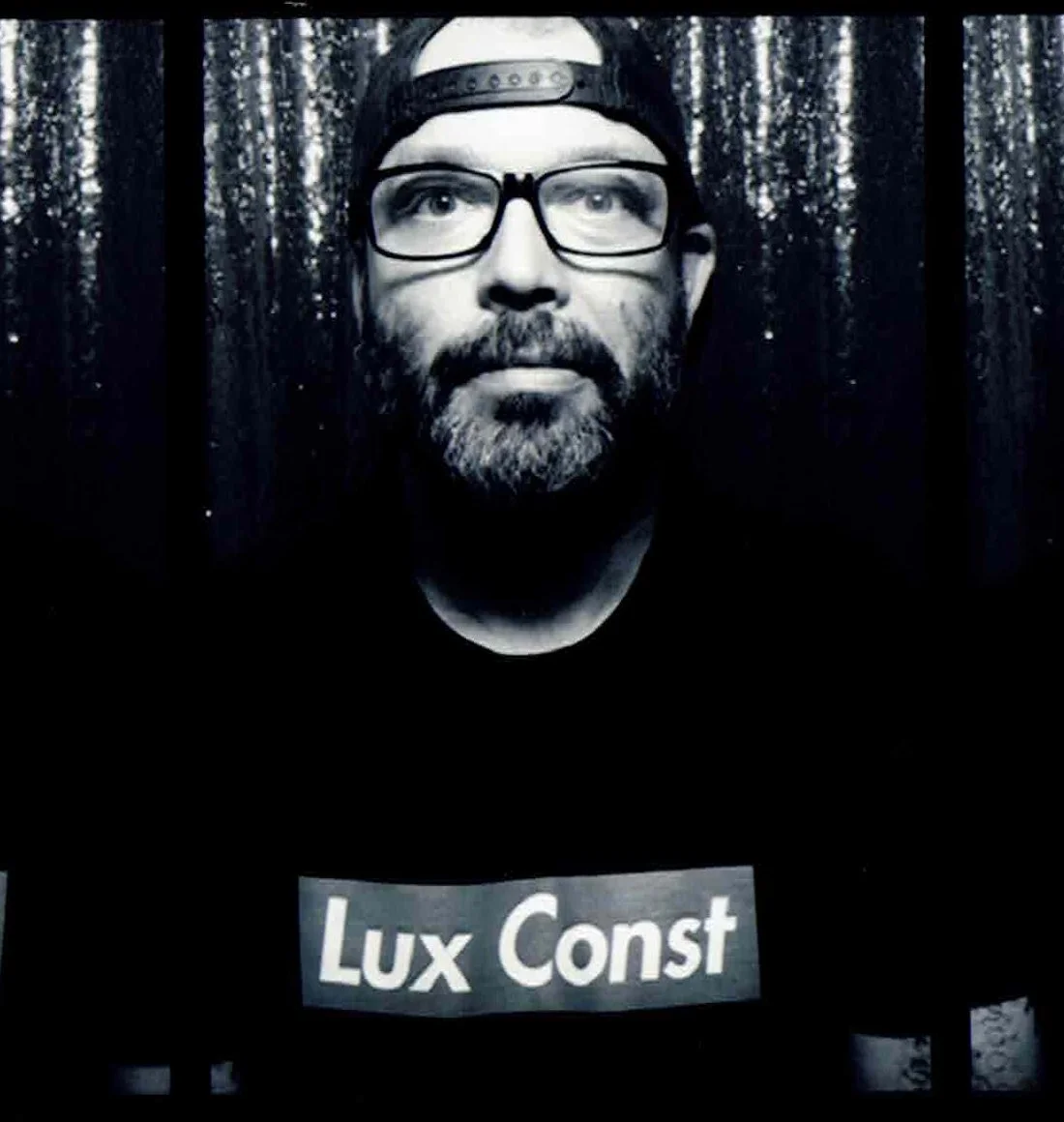 A young man with glasses and a beard wearing a backwards cap and a black T-shirt with "Lux Const" printed on it, standing in front of a dark, sparkly background.