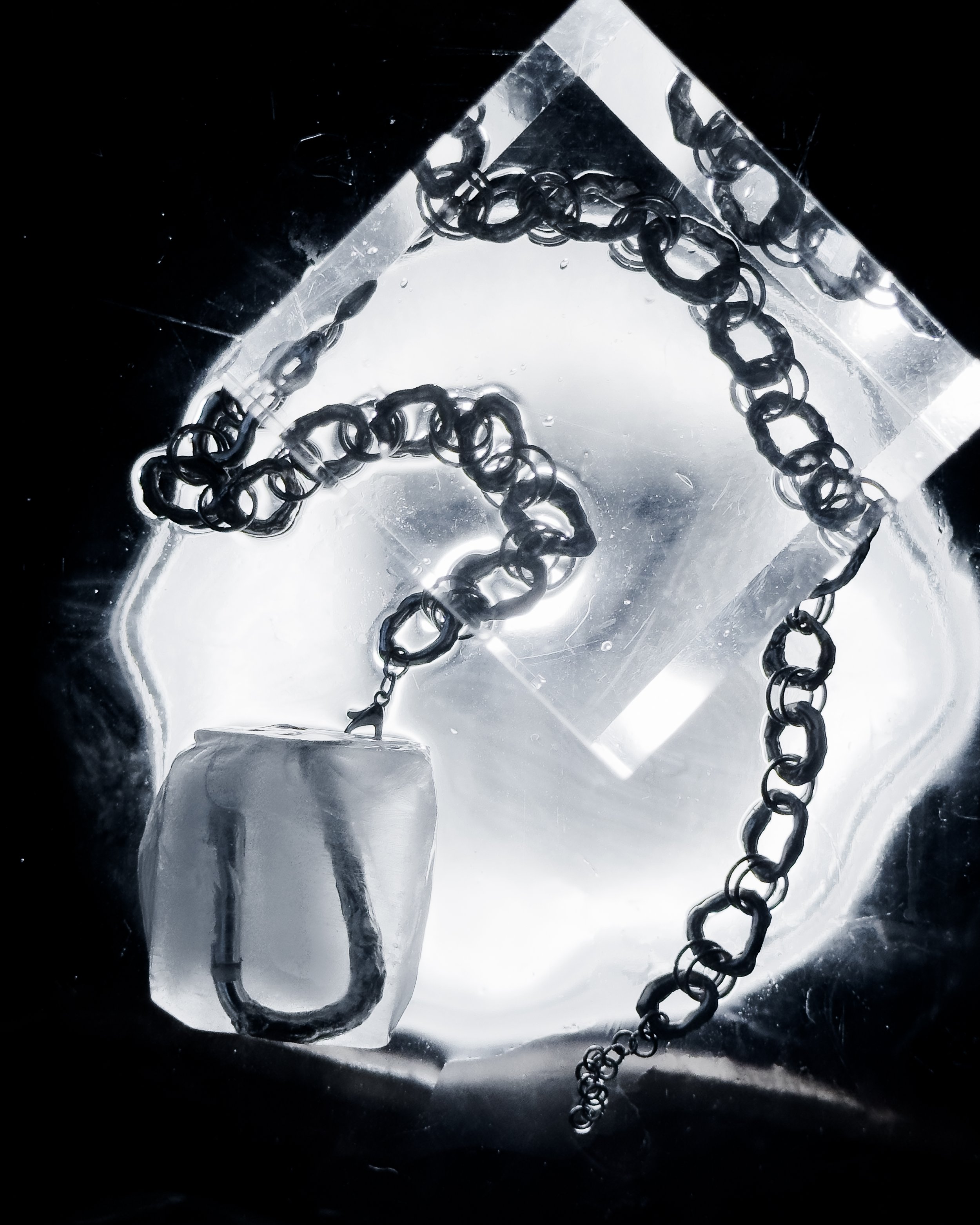 Close-up of a metal chain with a clasp inside a transparent ice cube, with another ice cube in the background, illuminated by a light source.
