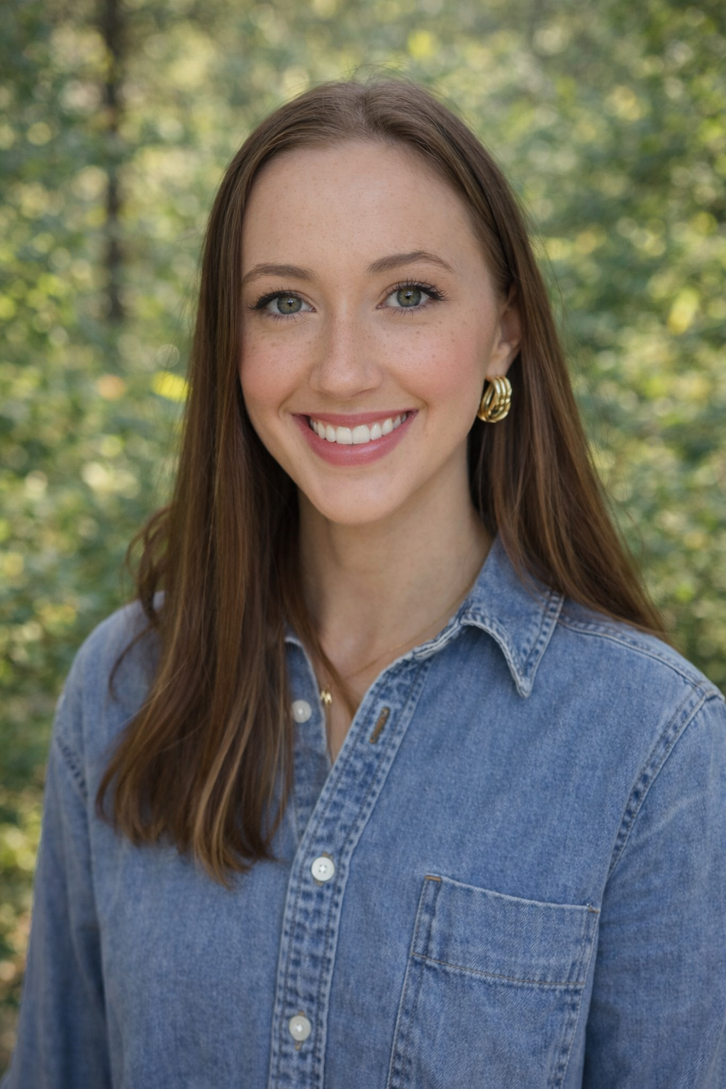 A young woman with long brown hair, blue eyes, and freckles, smiling and wearing a denim shirt and gold jewelry, standing in front of a white wall with green plant leaves behind her.
