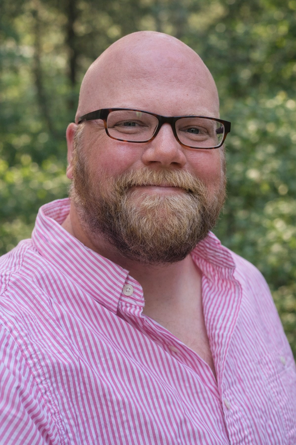 A man with glasses, a beard, and a bald head taking a selfie outdoors in a wooded area with trees and grass.