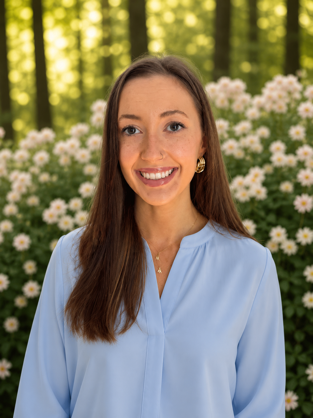 A young woman with long brown hair, blue eyes, and freckles, smiling and wearing a denim shirt and gold jewelry, standing in front of a white wall with green plant leaves behind her.