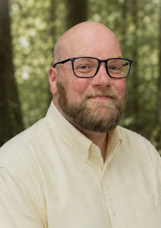 A man with glasses, a beard, and a bald head taking a selfie outdoors in a wooded area with trees and grass.