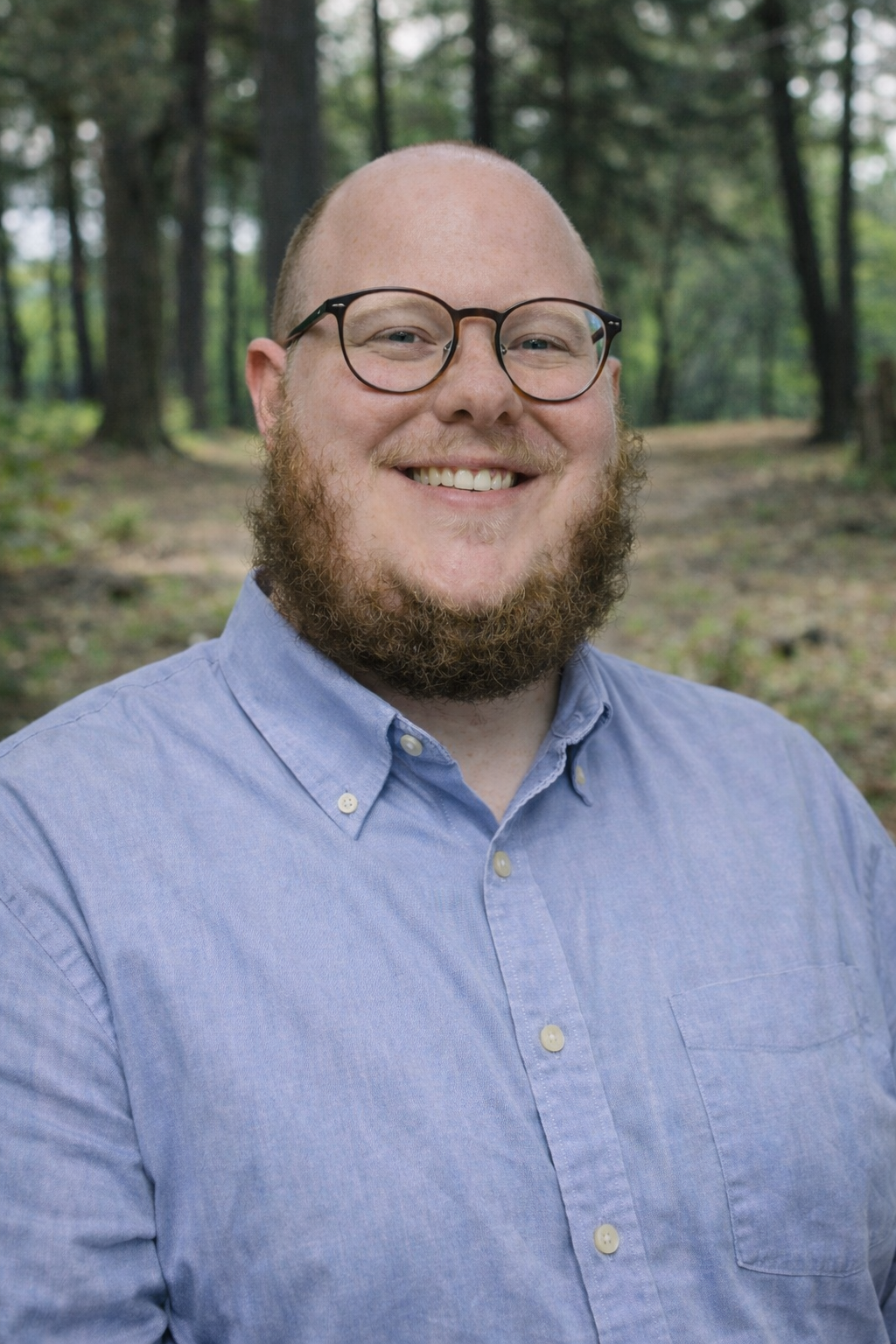 A smiling man with a beard, glasses, wearing a light-colored dress shirt and a floral patterned tie, standing against a plain white wall.