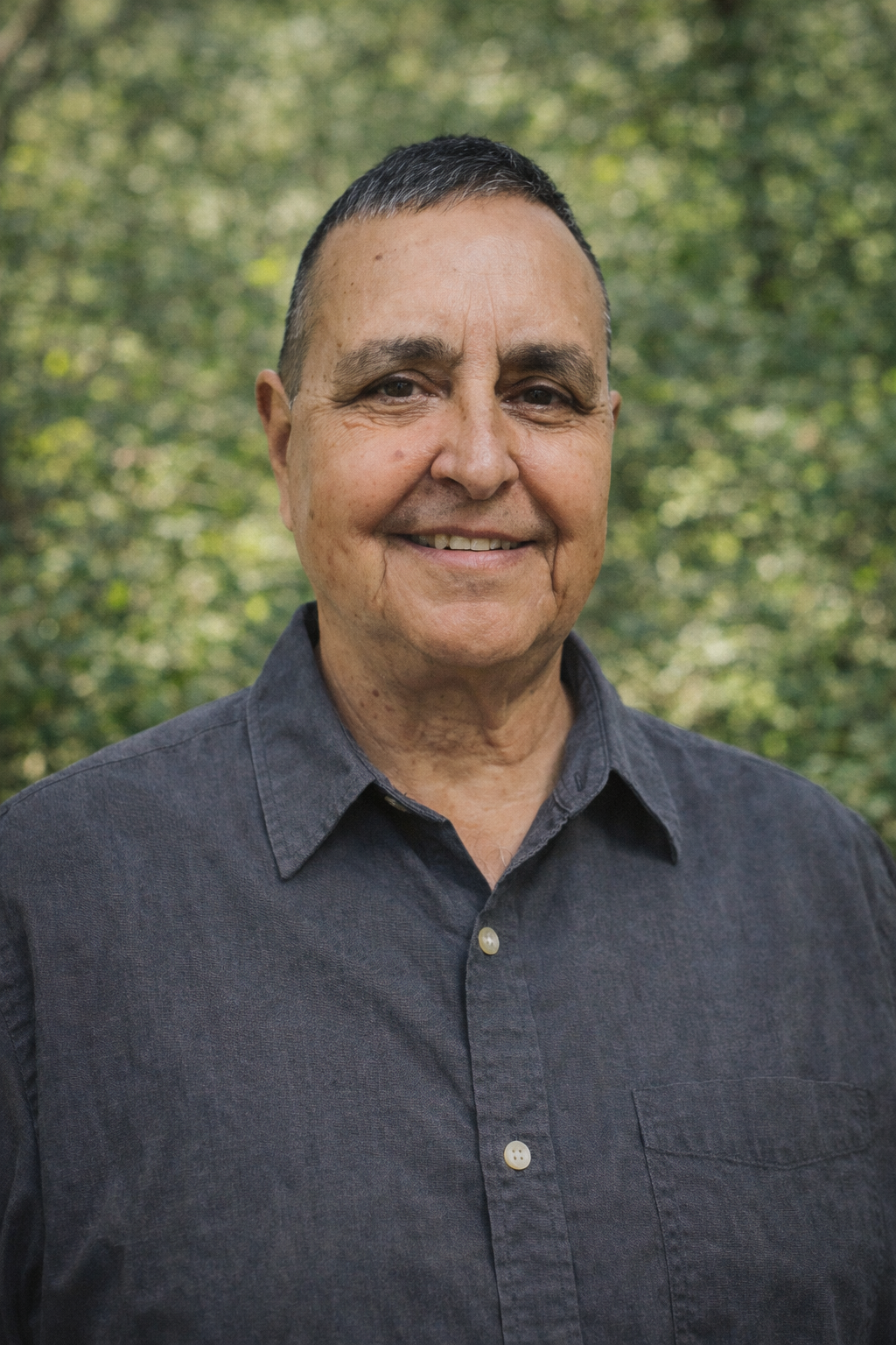Portrait of a smiling man with short dark hair, wearing a white button-up shirt, standing against a plain light gray background.