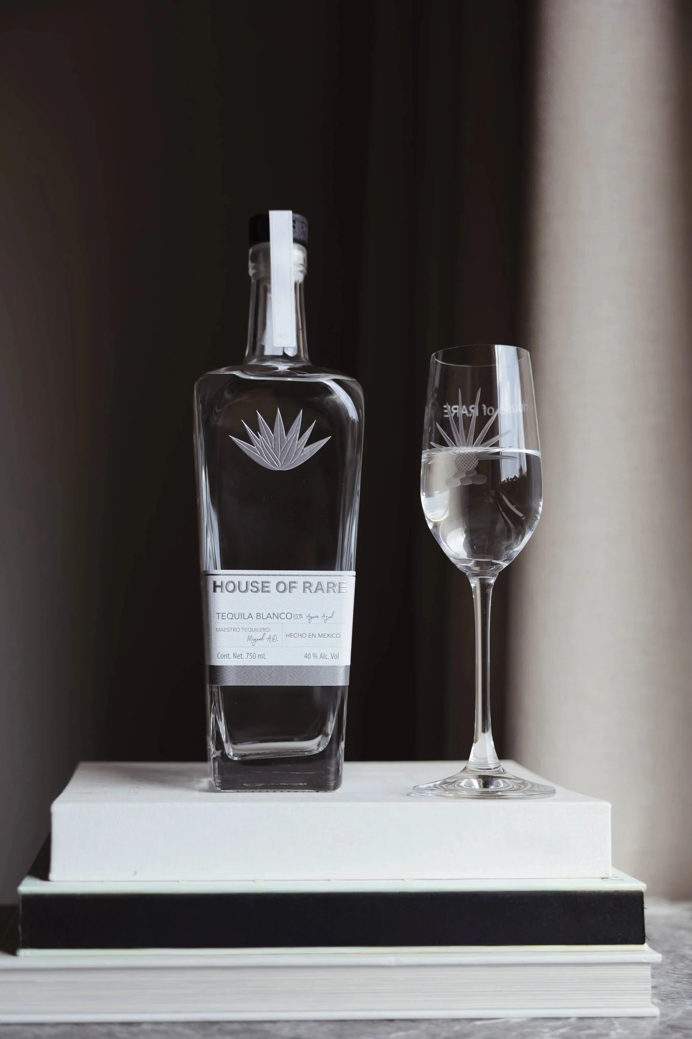 A bottle of House of Rare tequila blanco with a label, placed next to a glass of tequila on a white book and black hardcover book, set against a dark background with a light curtain to the right.