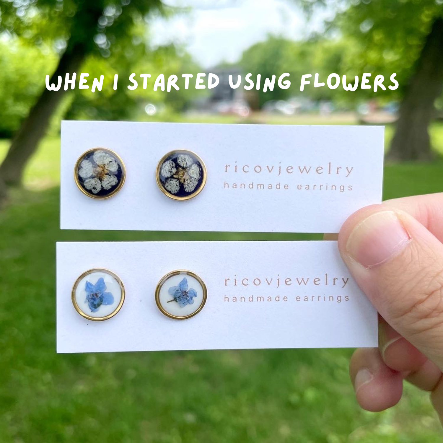 Hand holding two cards displaying pairs of handmade earrings with flower designs, one black with white flowers and gold accents, the other light blue with darker blue flowers, outdoors with green trees in the background.