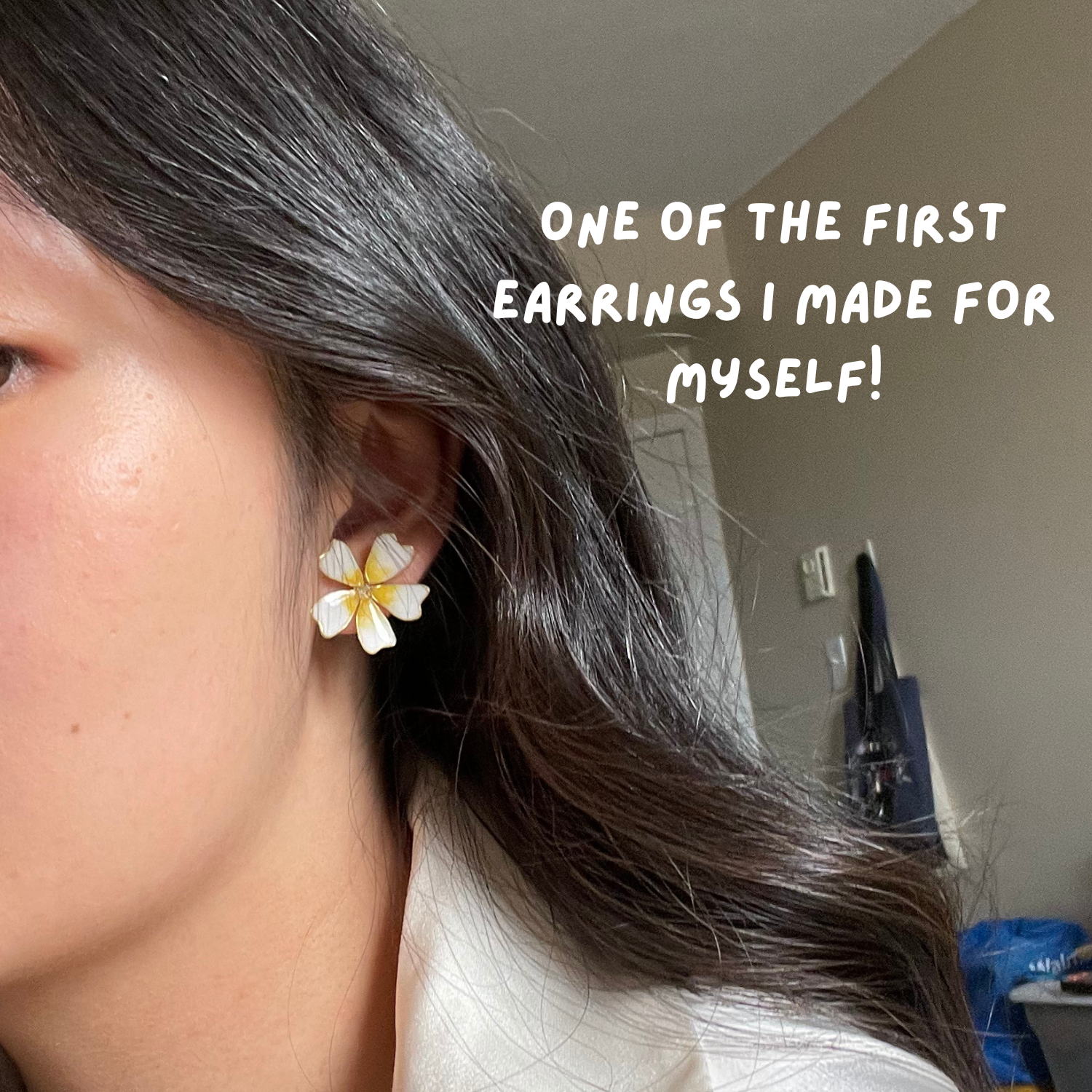 Close-up of a woman's ear wearing a flower-shaped earring with white and yellow petals, with text overlay saying 'One of the first earrings I made for myself!'