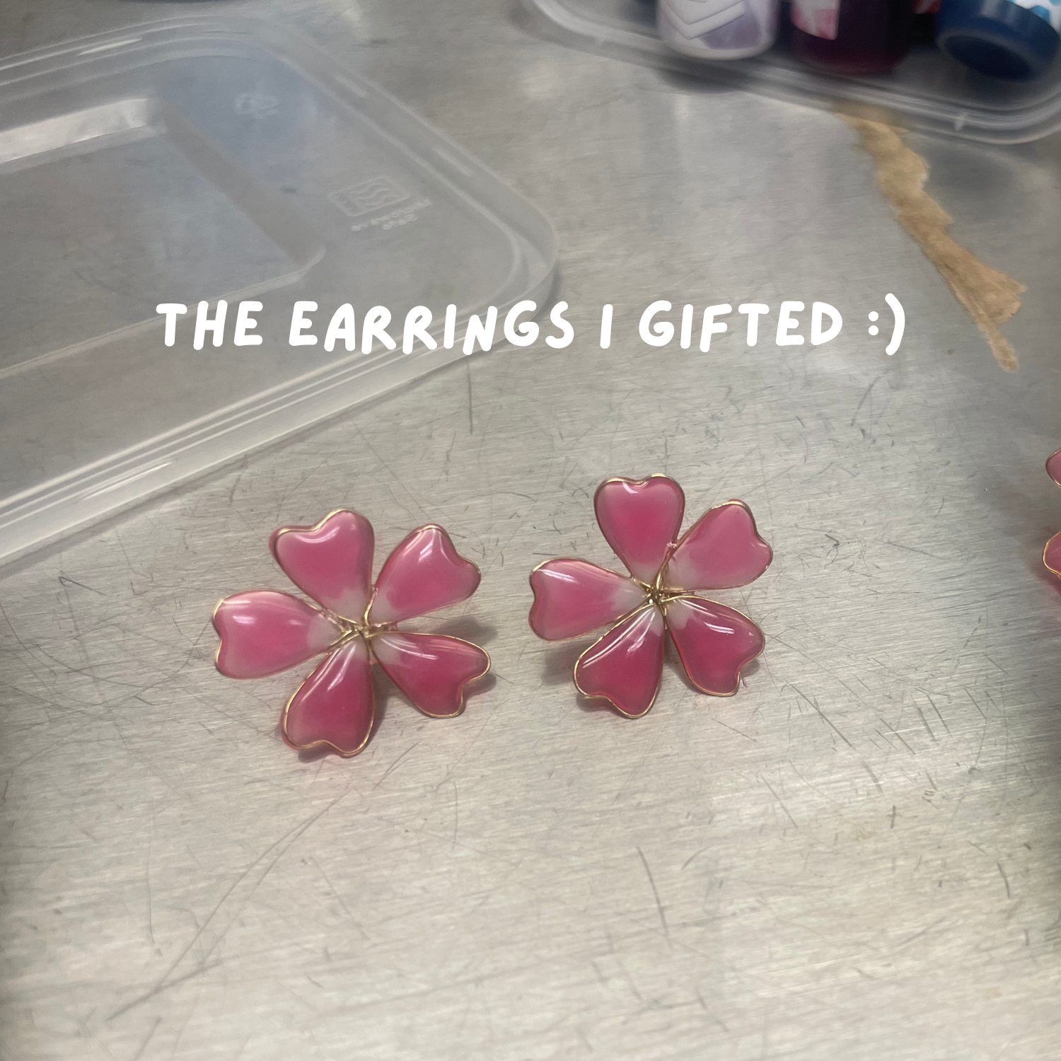 Pink flower-shaped earrings with gold edges placed on a metallic surface. Text overlay says "The earrings I gifted :)"