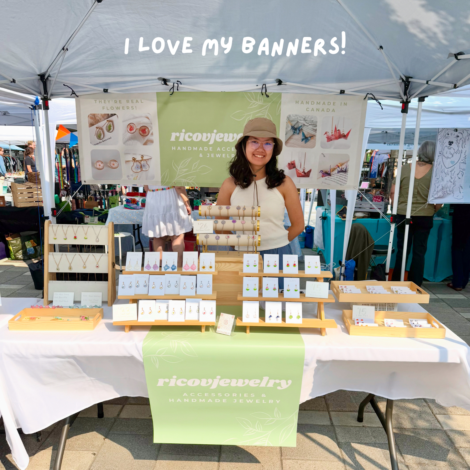 A woman standing behind a jewelry stall at an outdoor market, with handmade earrings and necklaces displayed on wooden racks. A green sign behind her reads 'ricojewelry' and 'handmade accessories & jewelry.' She is smiling, wearing glasses, a tan hat, and a sleeveless cream top.