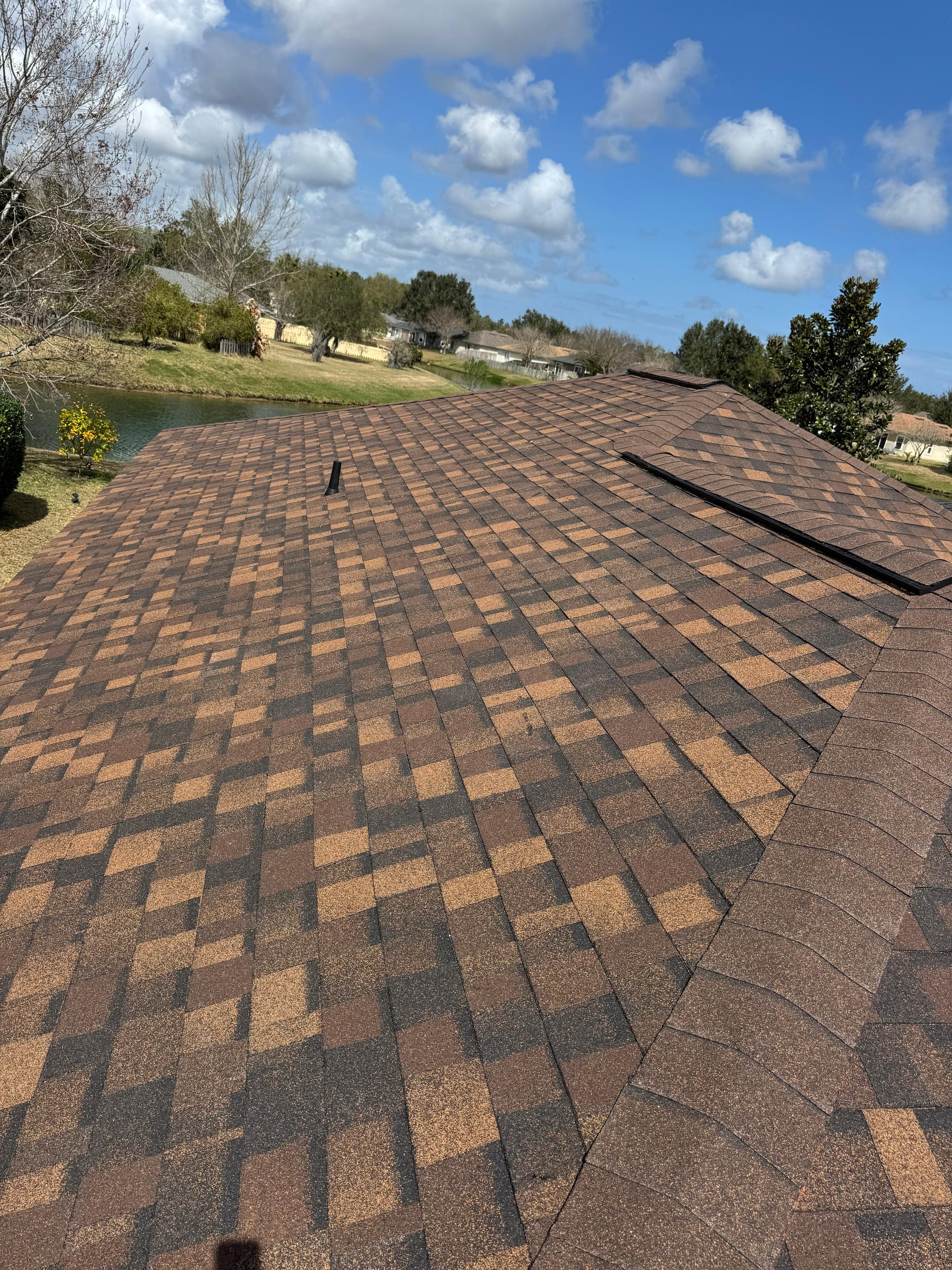 View of a house roof with asphalt shingles in shades of brown, blue sky with clouds, trees, and a pond landscape in the background.