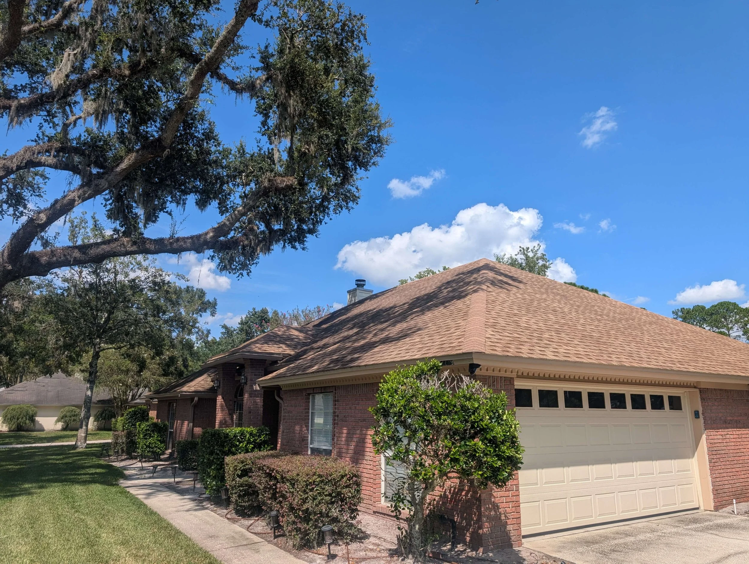 A suburban house with a brick exterior, tan garage door, and a front yard with trimmed bushes and trees under a clear blue sky.