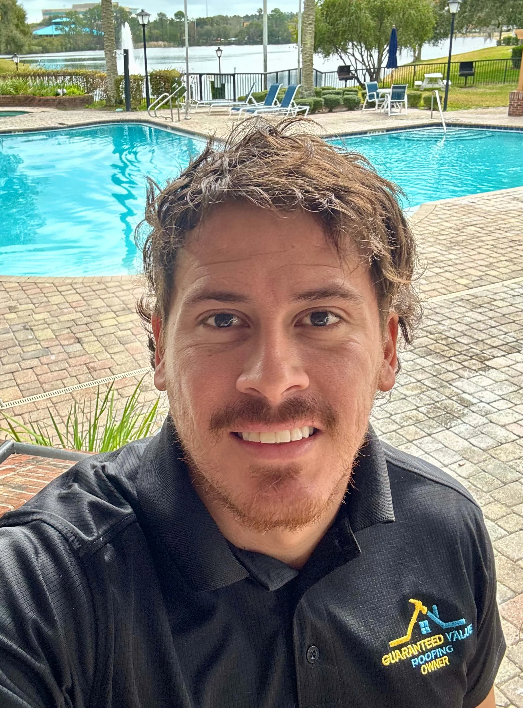 Selfie of a man with curly brown hair and a beard, smiling, wearing a black polo shirt with a logo, by a swimming pool with lounge chairs, umbrellas, trees, and a lake in the background.
