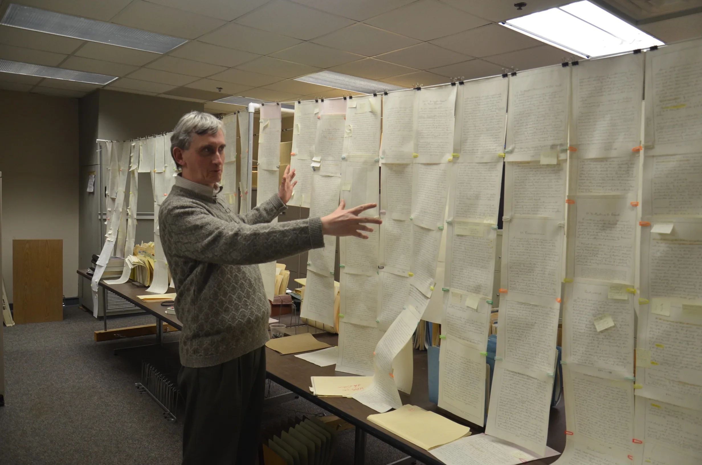 Man examining numerous handwritten or typed documents pinned on a large display board in a room, likely an archive or research facility.