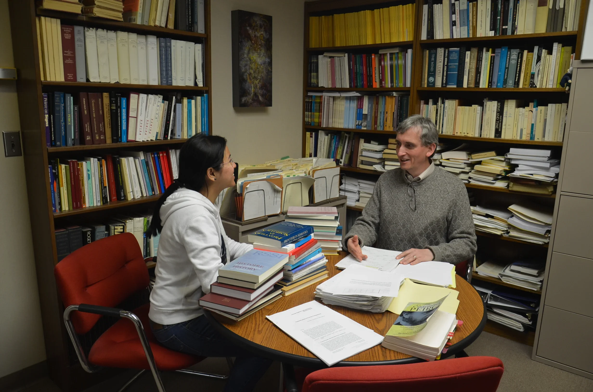 A woman and a man sitting across from each other at a round wooden table in an office. The table is covered with numerous books, papers, and documents. Behind them are shelves filled with books and files, and an abstract painting hangs on the wall.