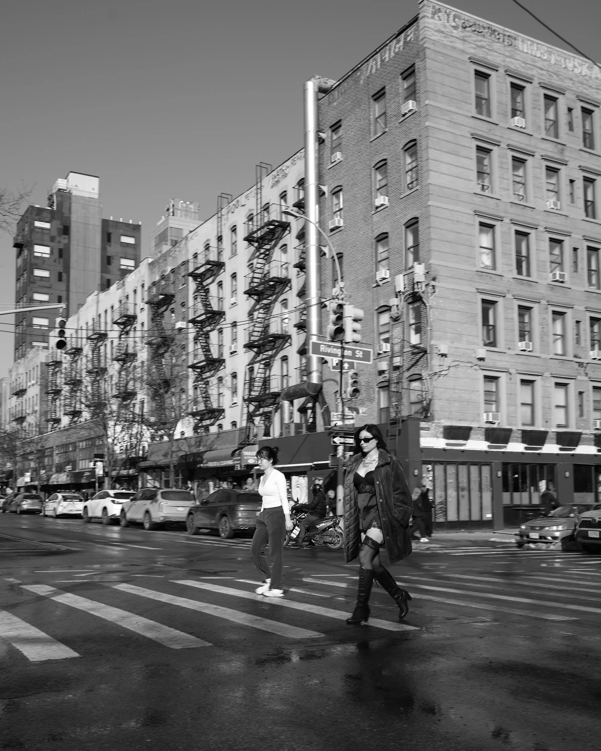 Two women crossing a city street, one in a white top and pants, the other in a black coat and thigh-high boots, with cars parked along the street and a multi-story building in the background.