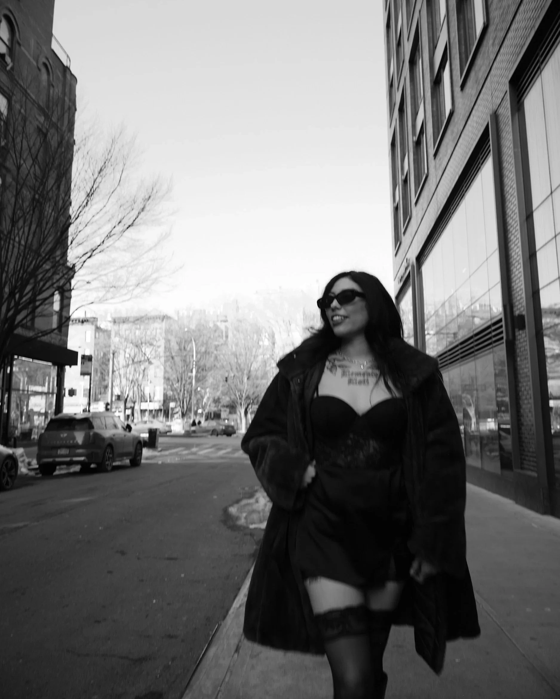 A woman in sunglasses and a strapless dress walking down a city sidewalk, smiling, with bare trees and buildings in the background in black and white.