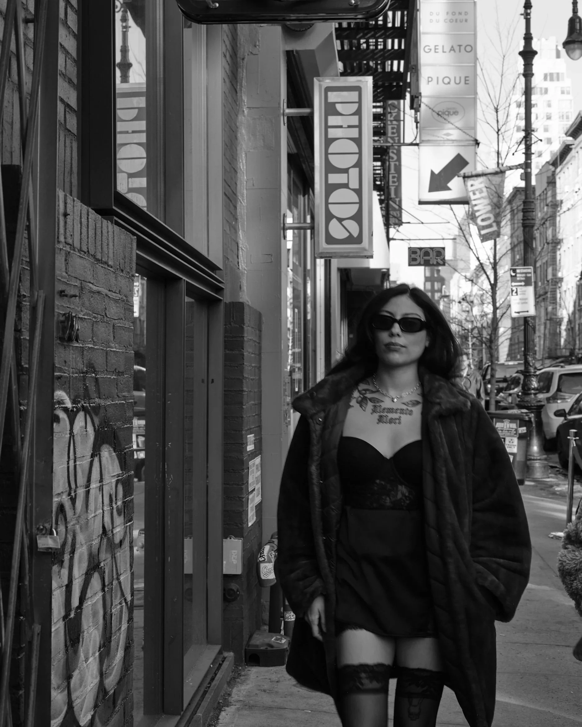 A woman wearing sunglasses, a black dress, and a coat, walking on an urban sidewalk in front of a store with signs for gelato and pizza. She has tattoos on her chest and thighs and long dark hair.