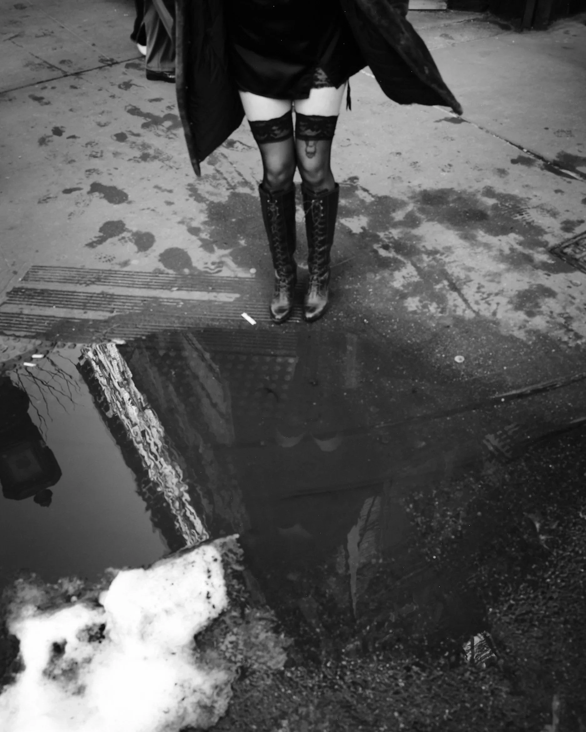 A person standing on a wet city street, wearing high boots, thigh highs, and dark clothing, with people's reflections visible in a puddle on the ground.
