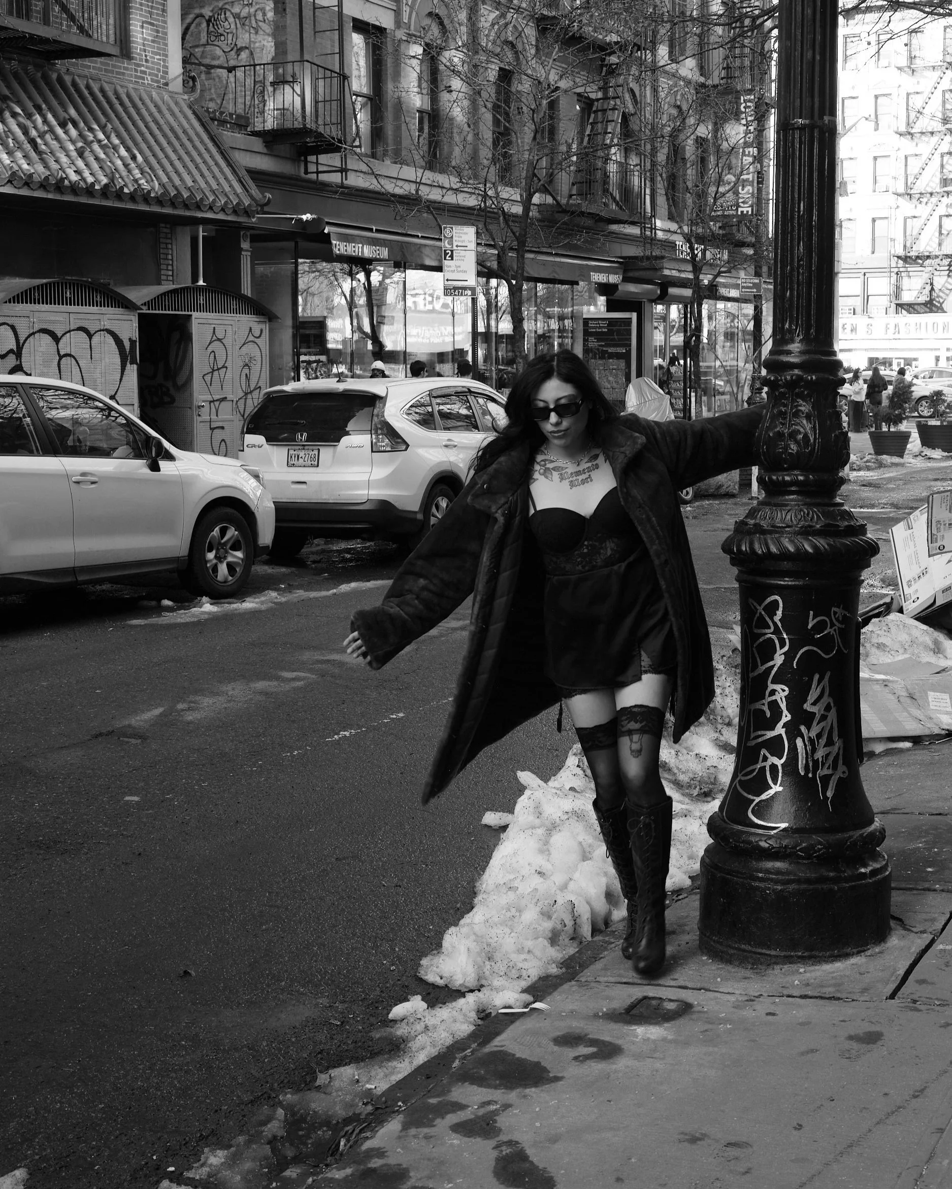 A woman with dark hair, sunglasses, and tattoos, wearing a short dress, knee-high stockings, and boots, leaning against a streetlamp on a city sidewalk with parked cars and buildings in the background.