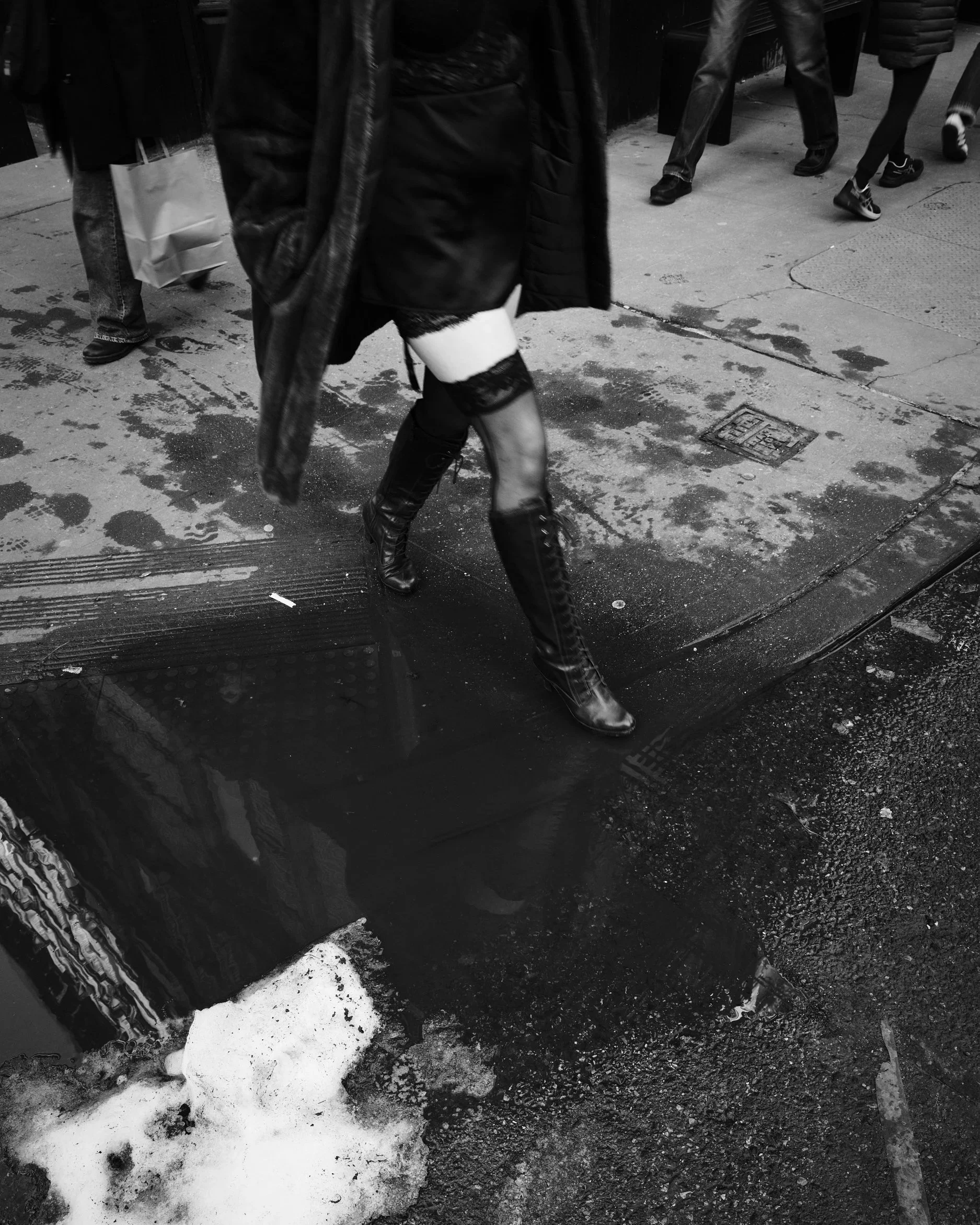A person walking on a city sidewalk wearing black thigh-high boots, a short skirt, and tights with lace tops, with other pedestrians and city street elements in the background.