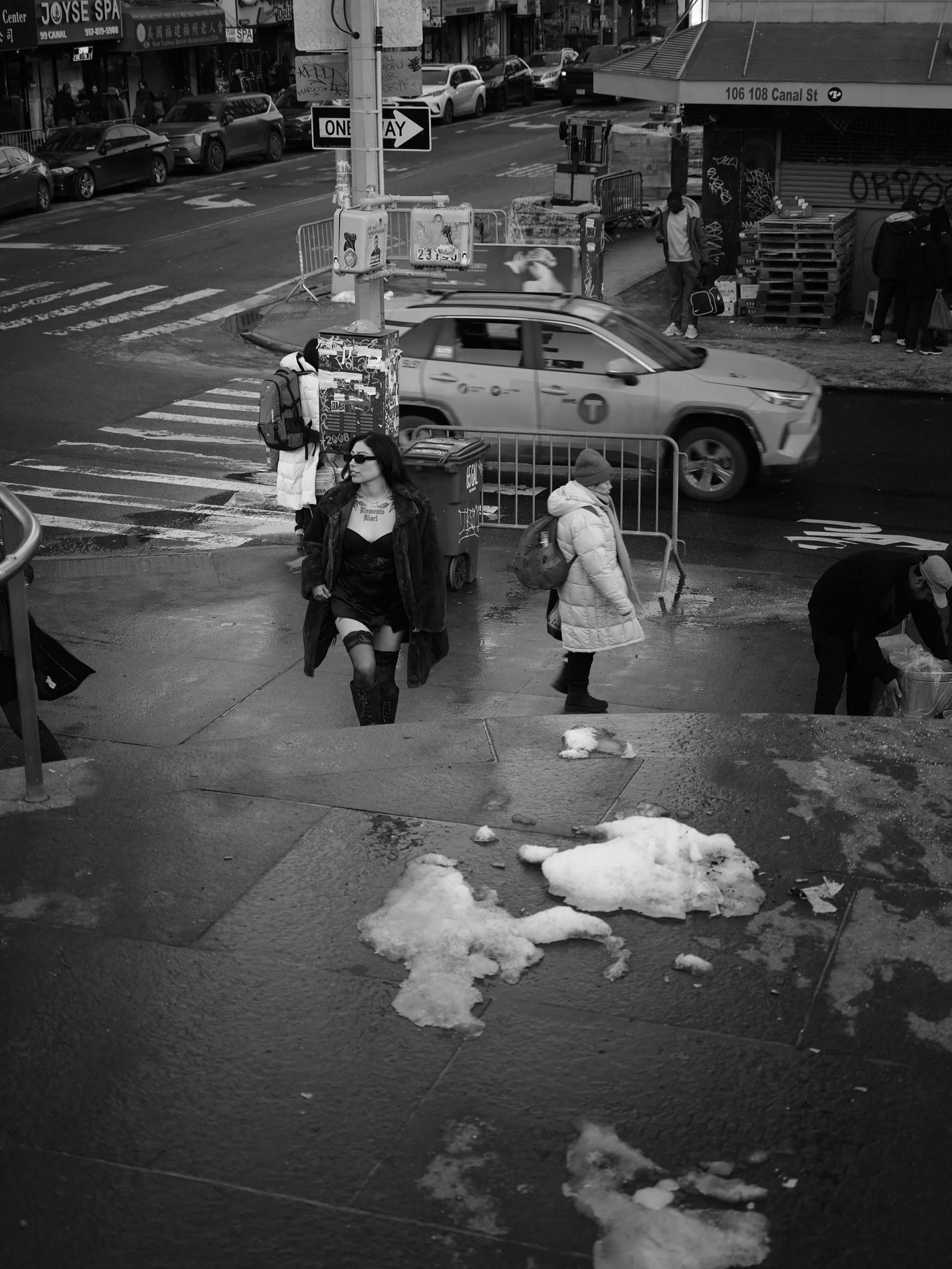 A busy urban street scene in black and white showing pedestrians crossing the street and walking on the sidewalk. There are cars and an SUV on the road, some graffiti on pillars, and snow patches on the sidewalk in winter weather.