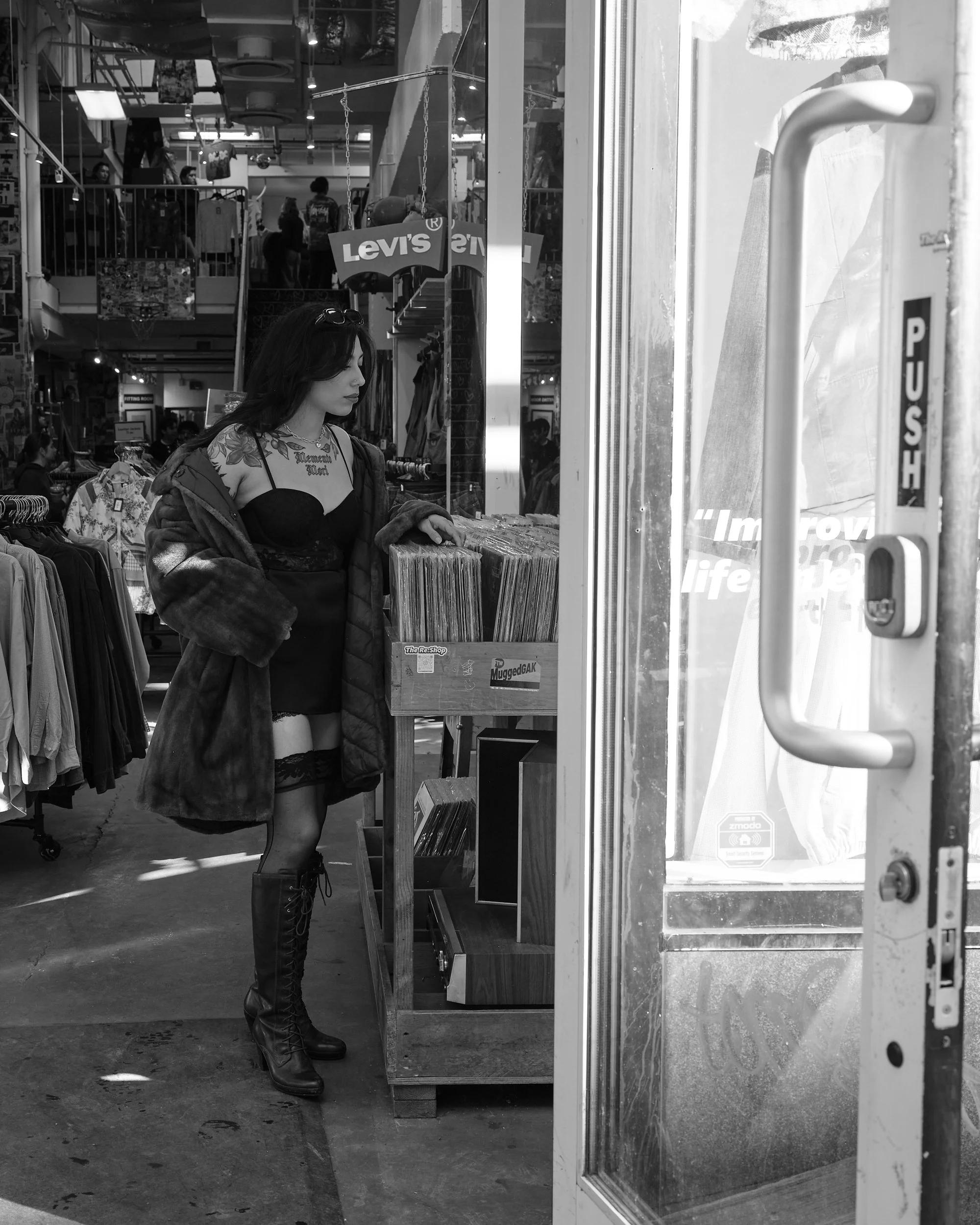 A woman shopping inside a store, looking at vinyl records. She is wearing a black dress, lace stockings, knee-high lace-up boots, and a jacket draped over her shoulders.