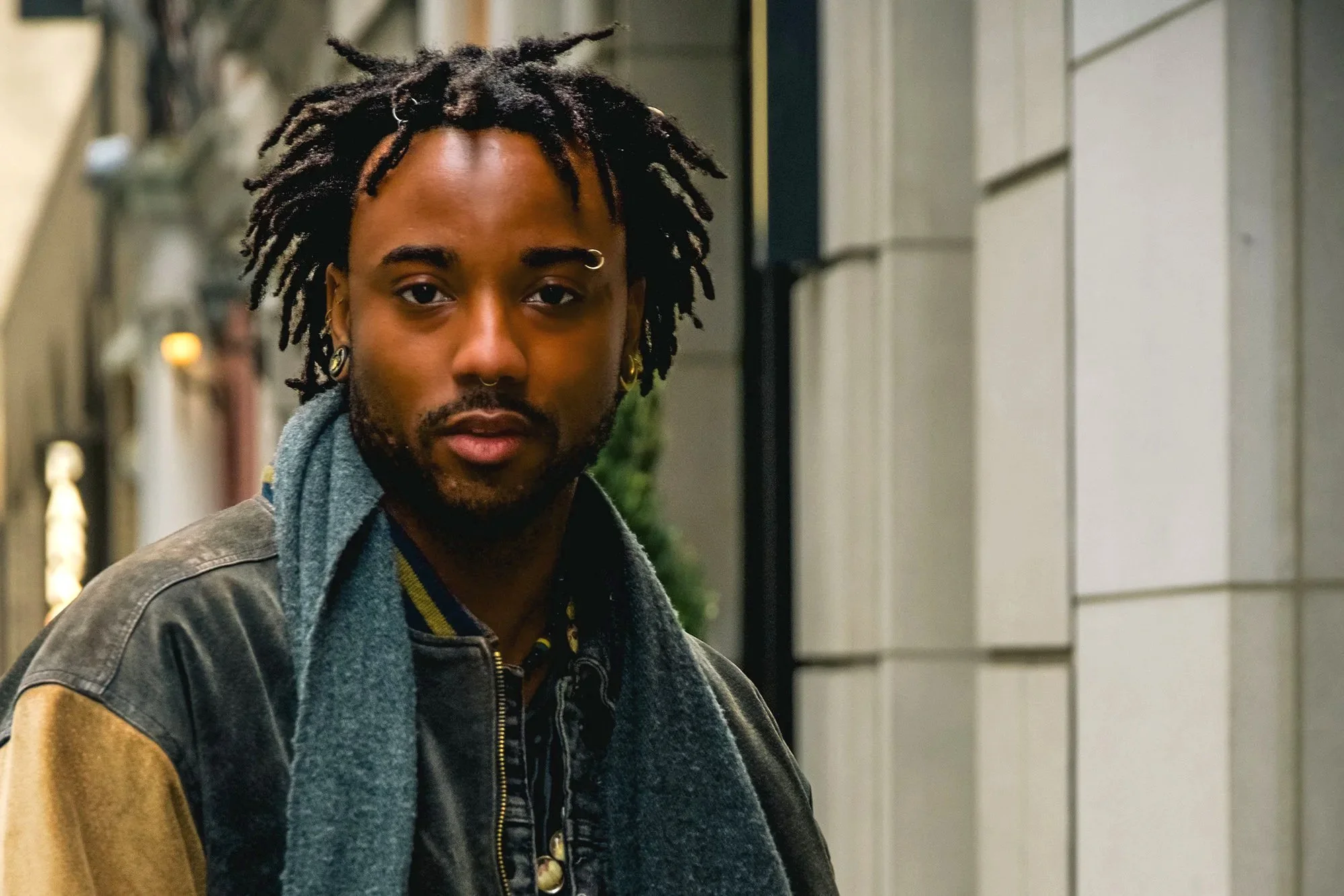 Professional headshot, photographed on location. A young man with dreadlocks, wearing earrings and a piercing, looking at the camera with a neutral expression, standing outdoors near a building.