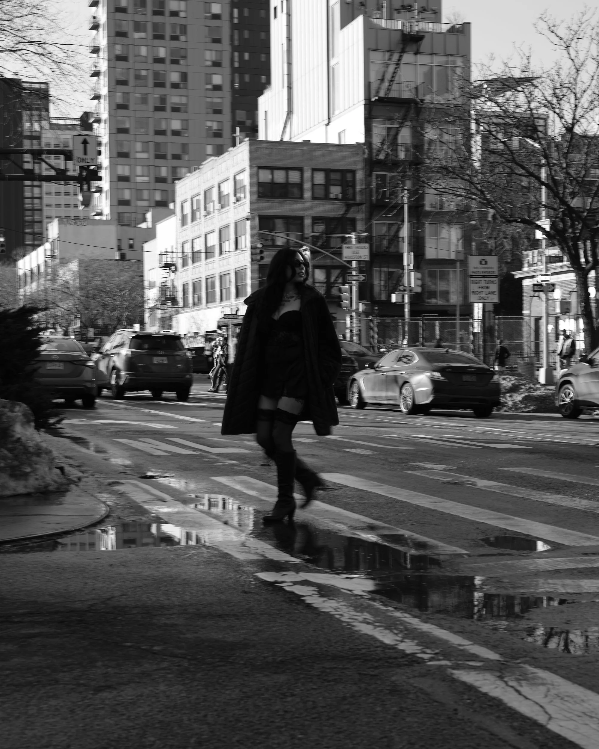 A woman dressed in black with thigh-high boots walking across a city crosswalk in black and white, with high-rise buildings, cars, and leafless trees in the background.