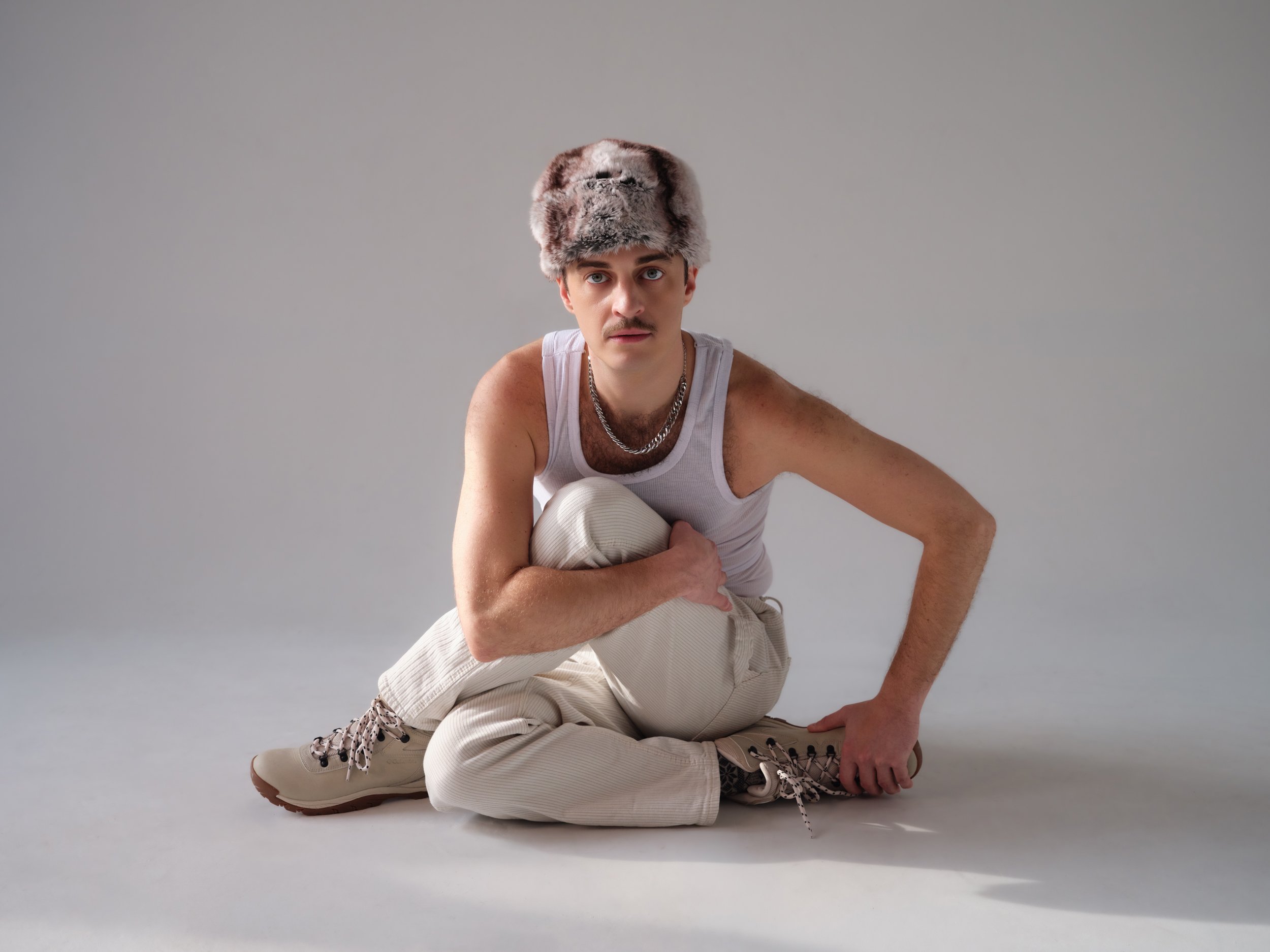 A young man with a mustache and light hair, wearing a faux fur hat, a white tank top, beige pants, gray boots, and a chain necklace, is sitting on the floor in a pose with one knee up and arms resting on his legs against a plain gray background.