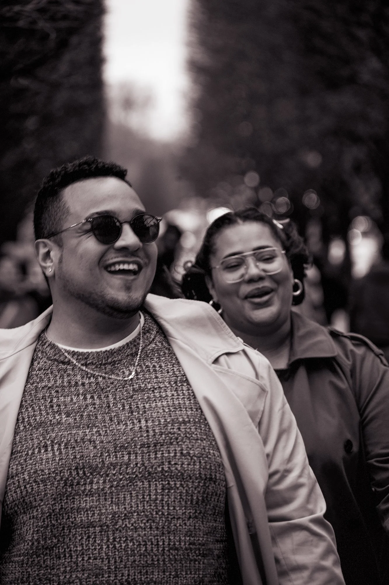 Two smiling people, a man wearing sunglasses and a woman with curly hair and glasses, outdoors with blurred trees in the background.
