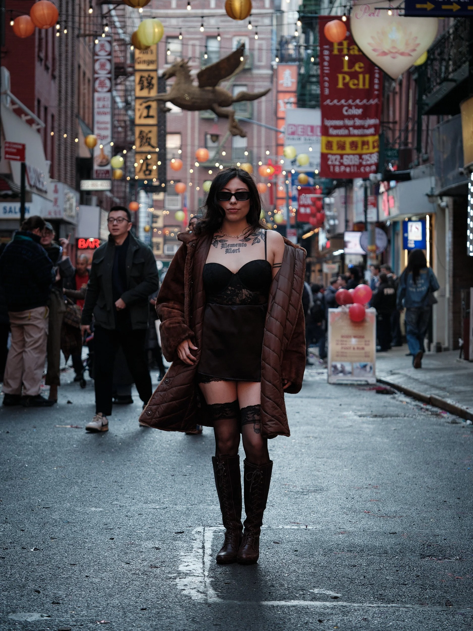 A woman wearing sunglasses, a black dress with lace details, thigh-high stockings, and knee-high boots walking down a busy street decorated with colorful lanterns and signs, with other pedestrians in the background.