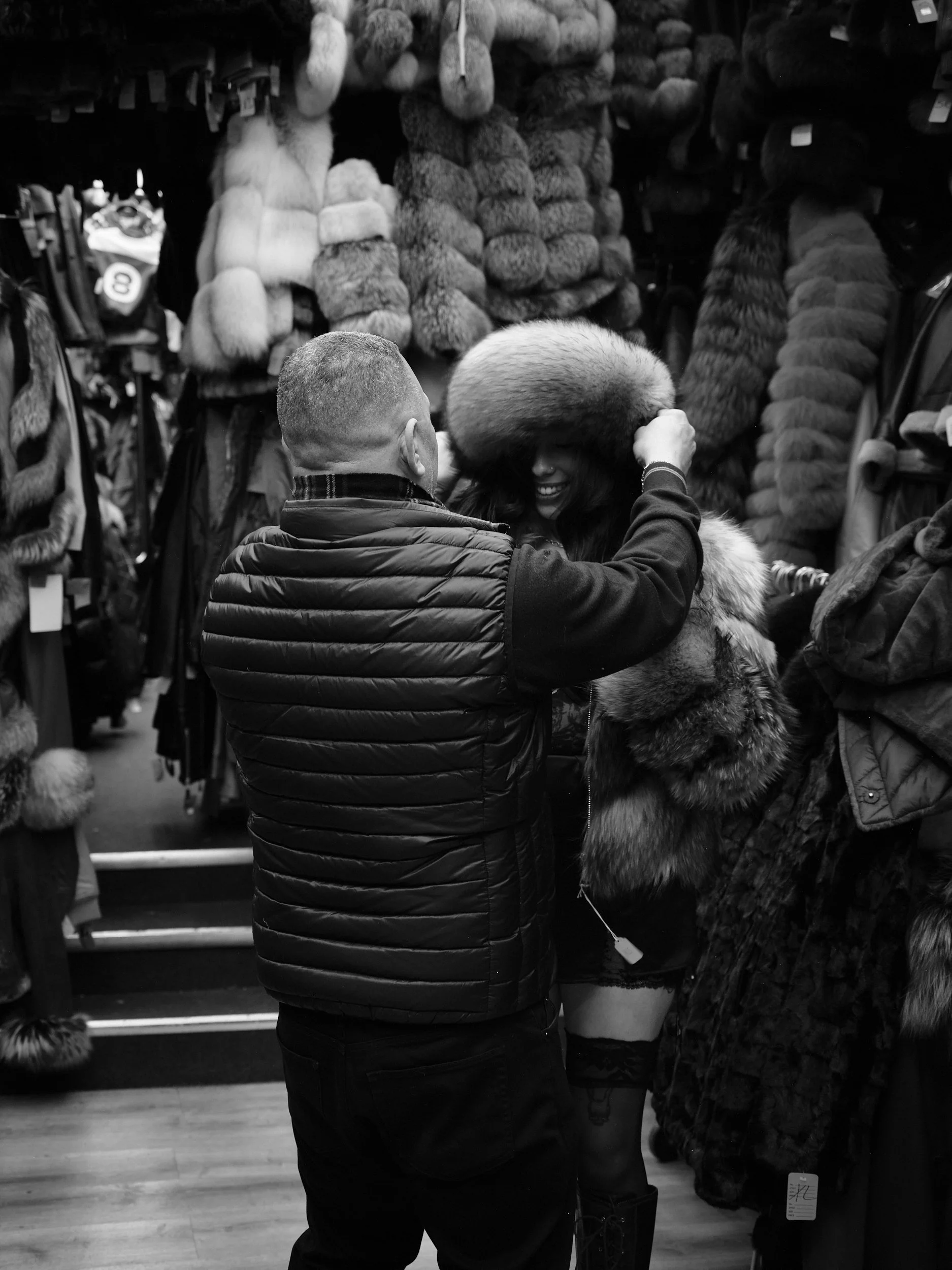 A man and woman shopping for fur coats at a store. The woman is trying on a large fur hat, and they are surrounded by various fur coats on display.