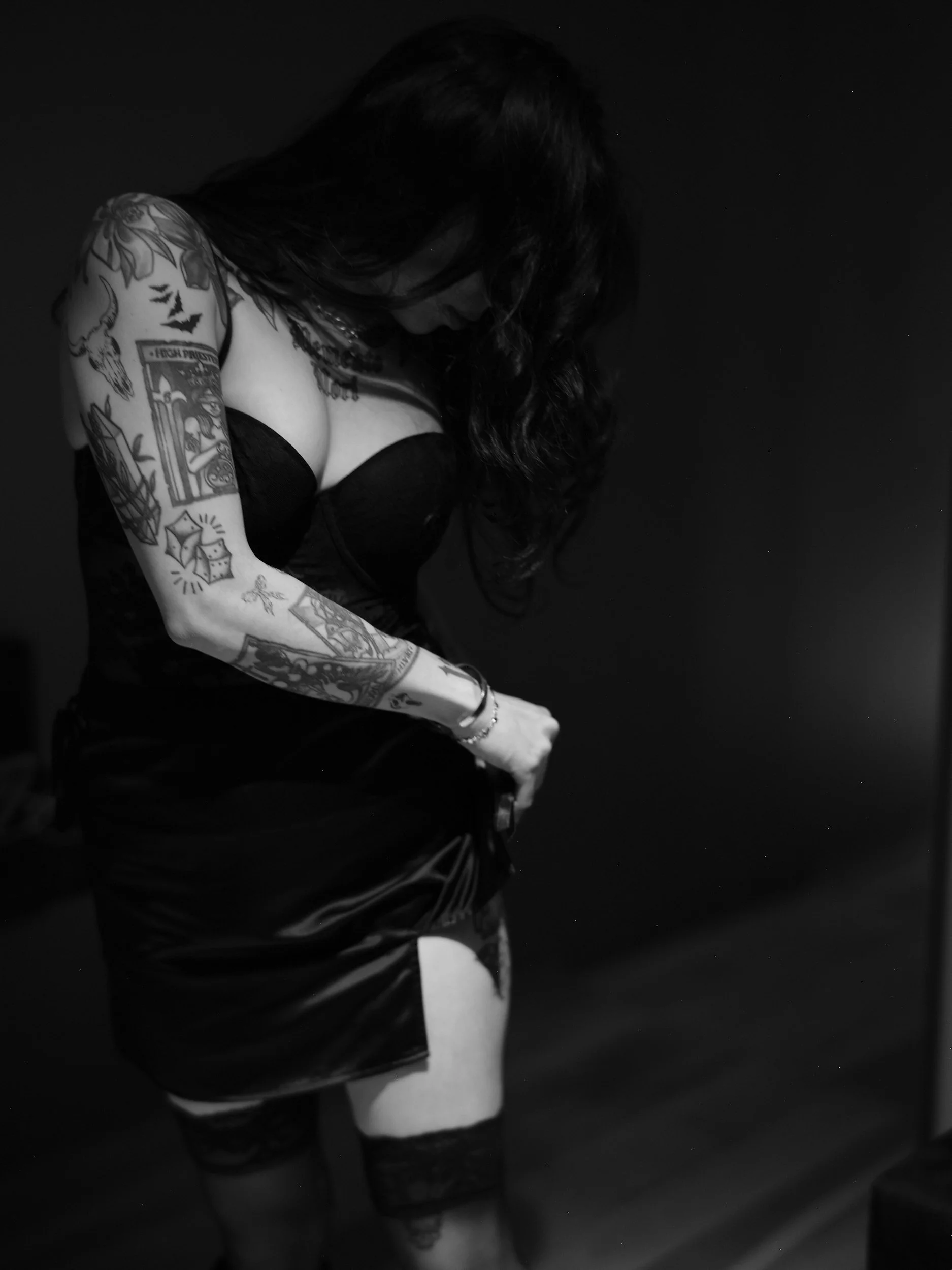 A woman with long dark hair and tattoos, wearing a black strapless top, black skirt, and thigh-high lace stockings, is adjusting her clothing against a dark background.