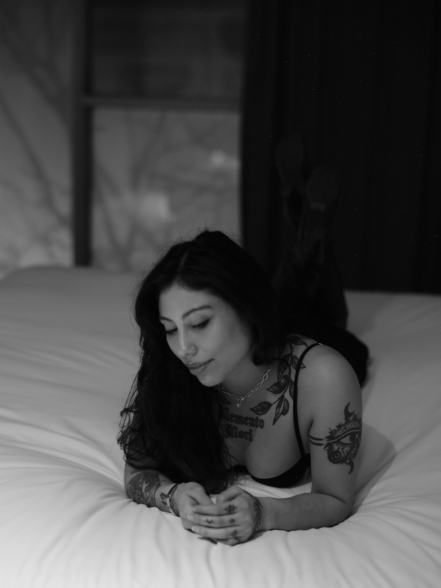 A young woman with tattoos and dark hair lying on a bed, looking at her phone, in a black and white photo.