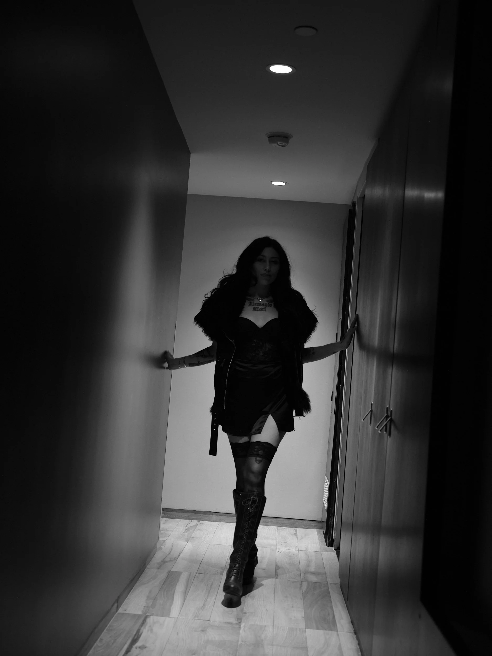 A woman walking through a narrow hallway, wearing a black mini skirt, lace stockings, and lace-up boots, with her hands touching the walls on either side.