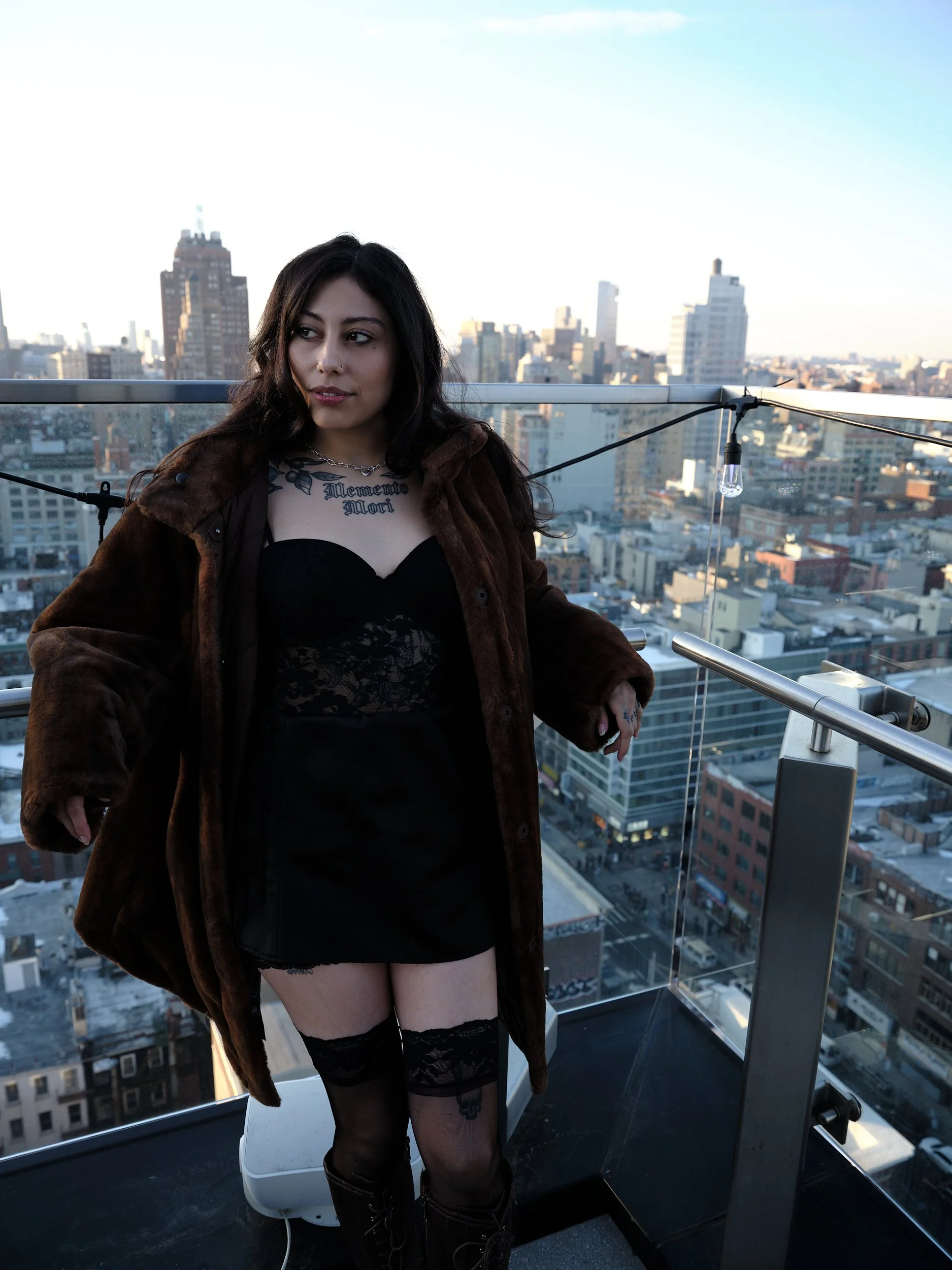 A young woman standing on a rooftop observation deck in a city, wearing a black dress, thigh-high stockings, and a brown fur coat, with city buildings in the background during daylight.