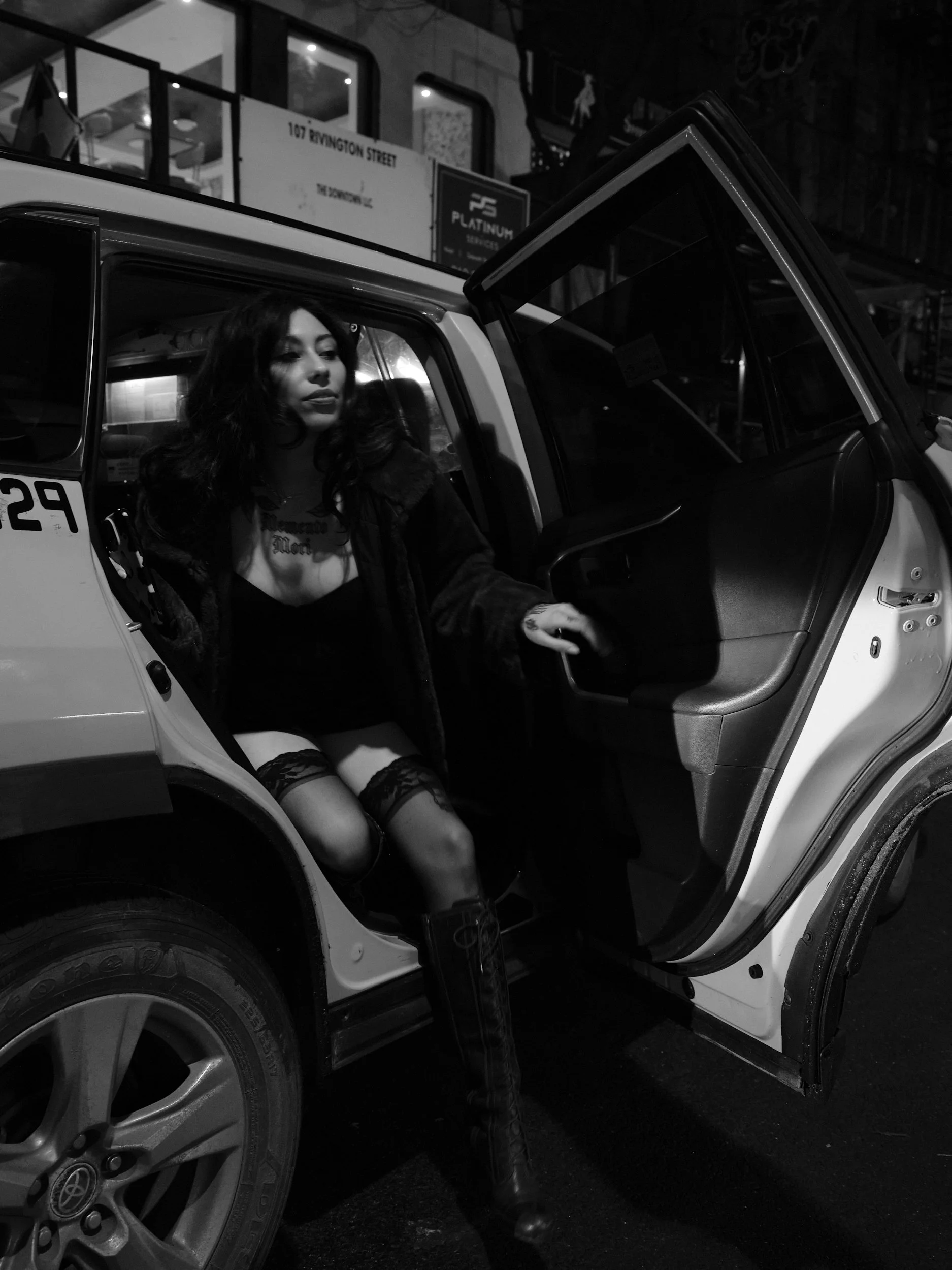 A woman in dark clothing, with stockings and lace, sitting inside a police car at night with the car door open.