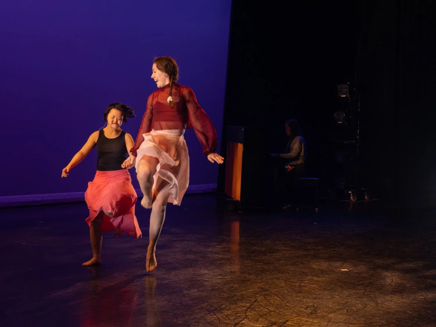 Two women dancing on stage under purple and orange lights, with one woman jumping in the air and the other smiling and moving.