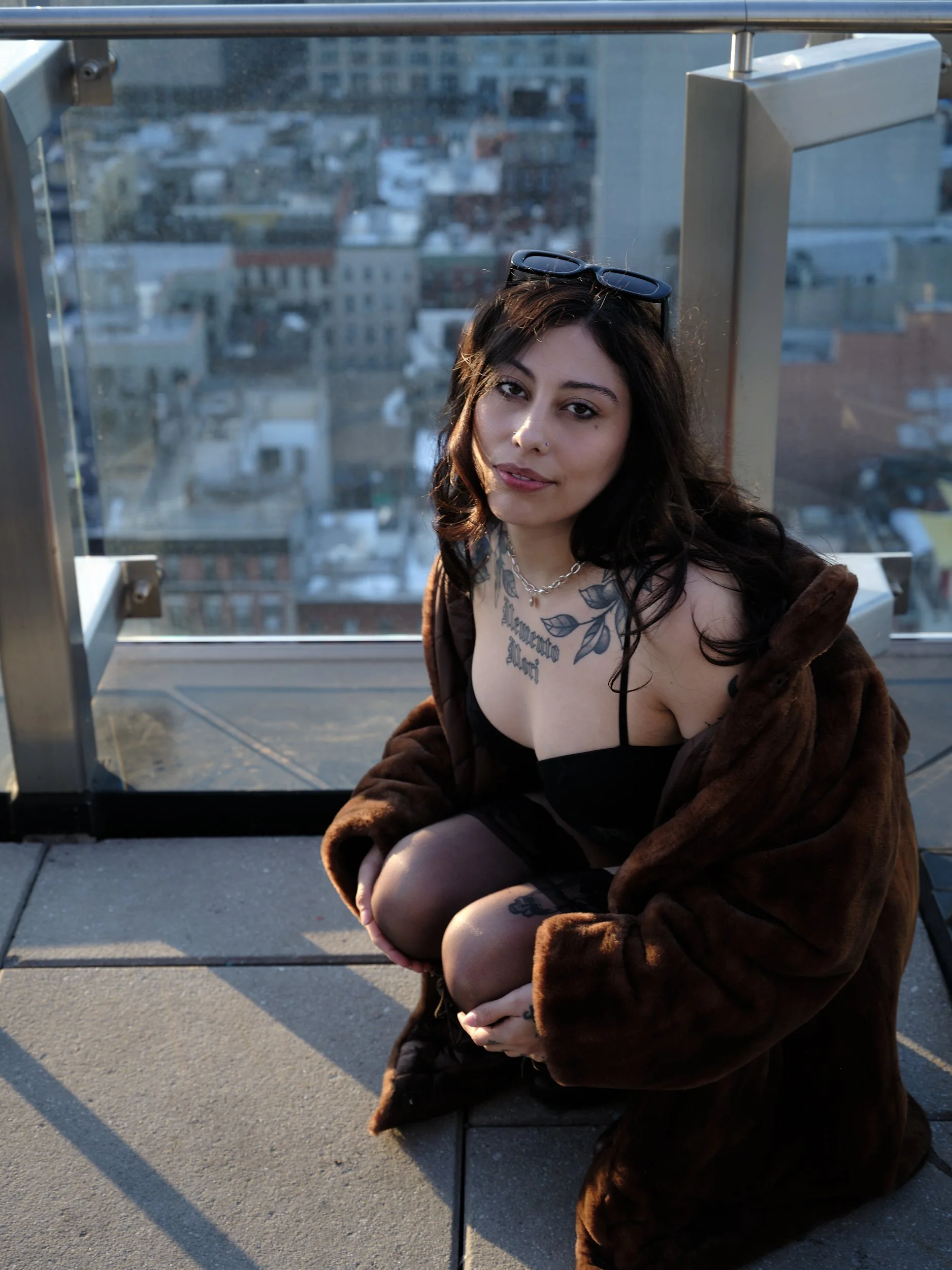 A woman with dark hair, wearing glasses on her head, a black top, and a brown fur coat, crouching on a rooftop balcony with a city view in the background, visible behind a glass railing.