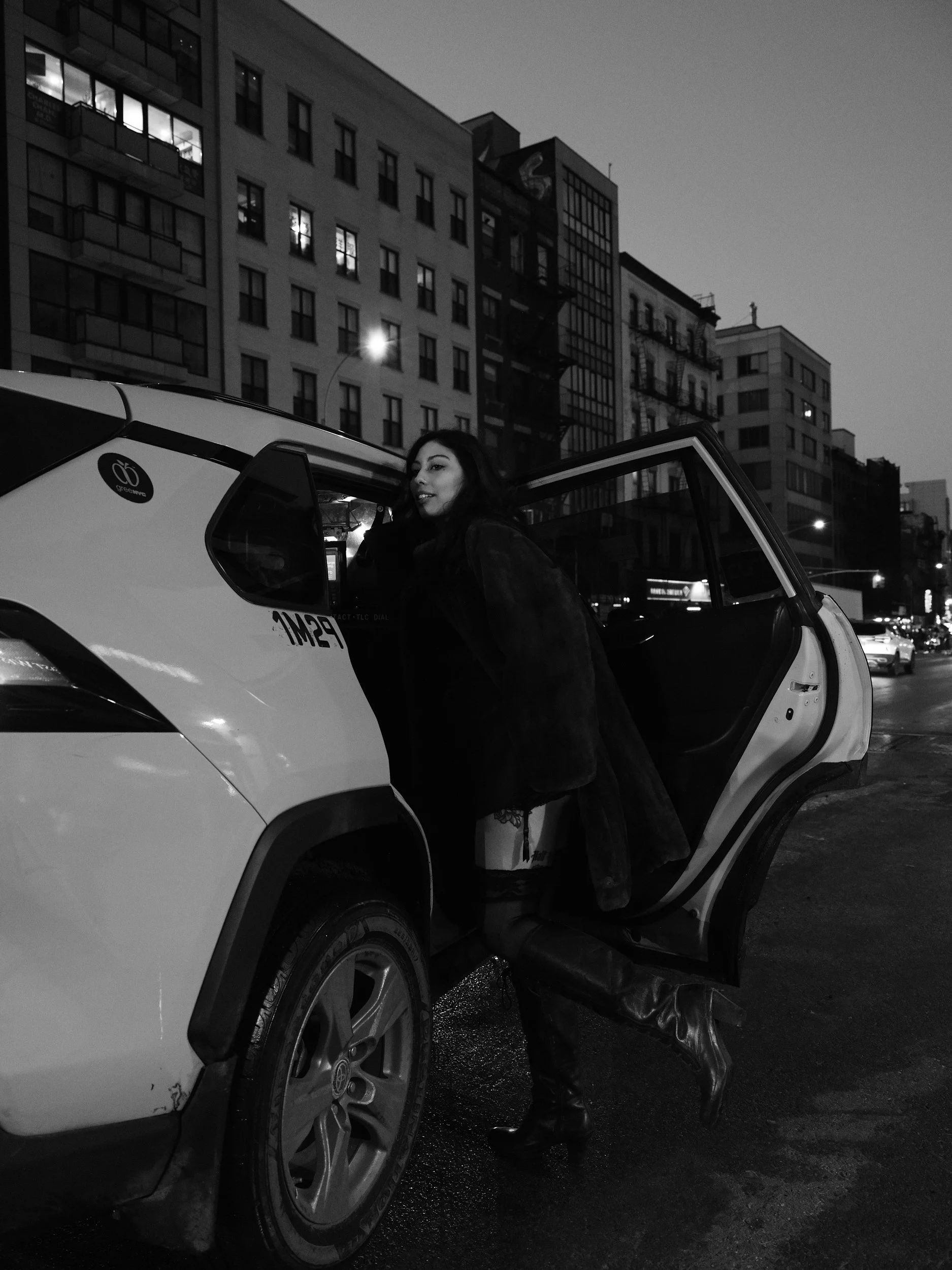 A woman with dark hair and dark clothing stepping out of a white SUV on a city street at dusk. The SUV's door is open, and the background shows tall buildings with lit windows.