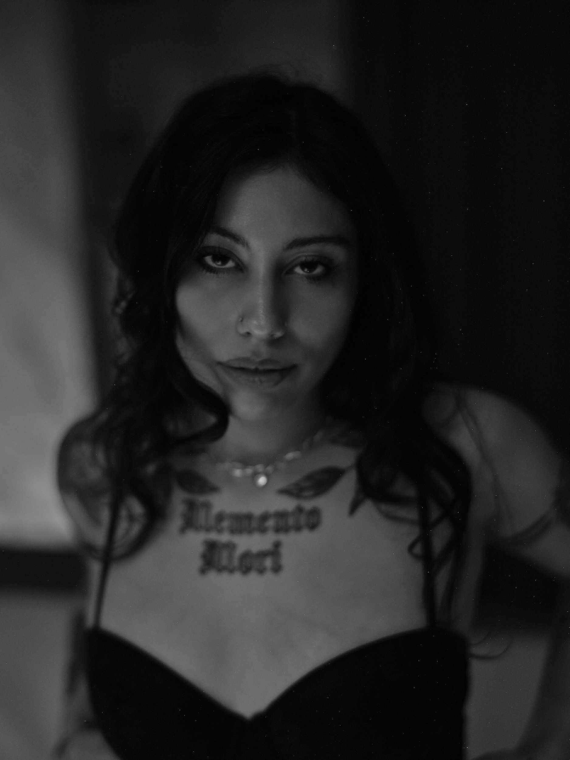 A woman with dark hair and tattoos, wearing a necklace and a top with spaghetti straps, looking directly at the camera in black and white.