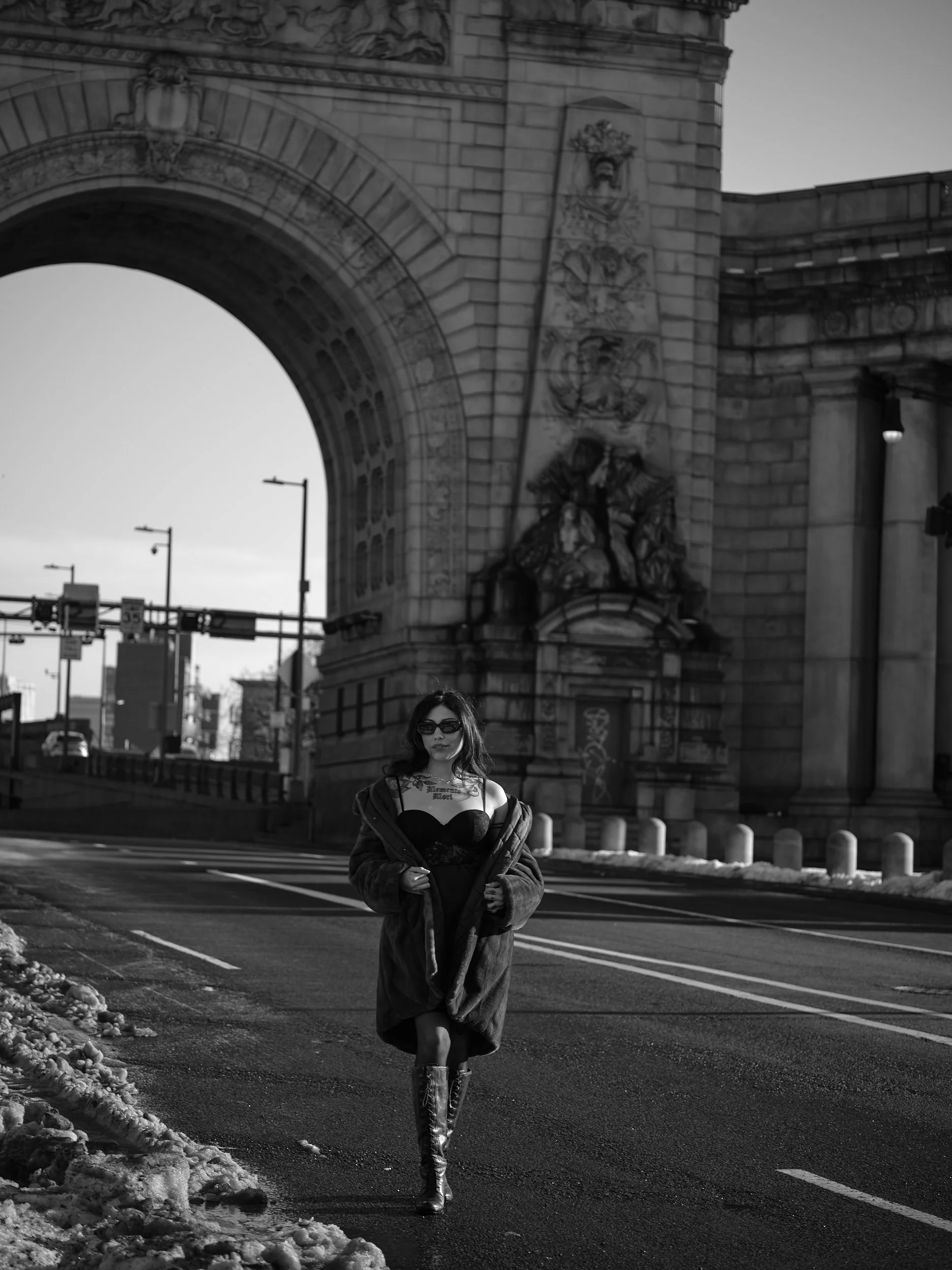A woman walking on a city street near a large stone archway, wearing sunglasses, a black dress, knee-high boots, and a coat, with graffiti on the wall behind her in black and white.