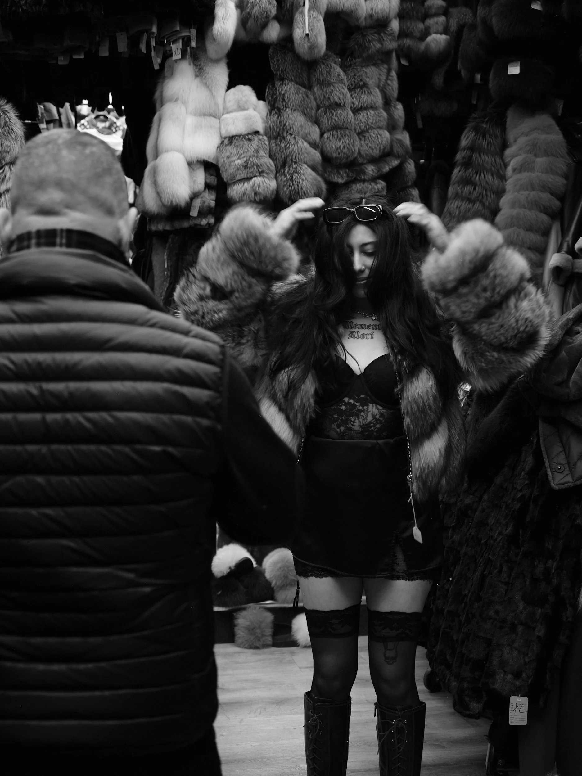 A woman trying on sunglasses in a fur coat store, surrounded by hanging fur coats, with two men in jackets looking at her.