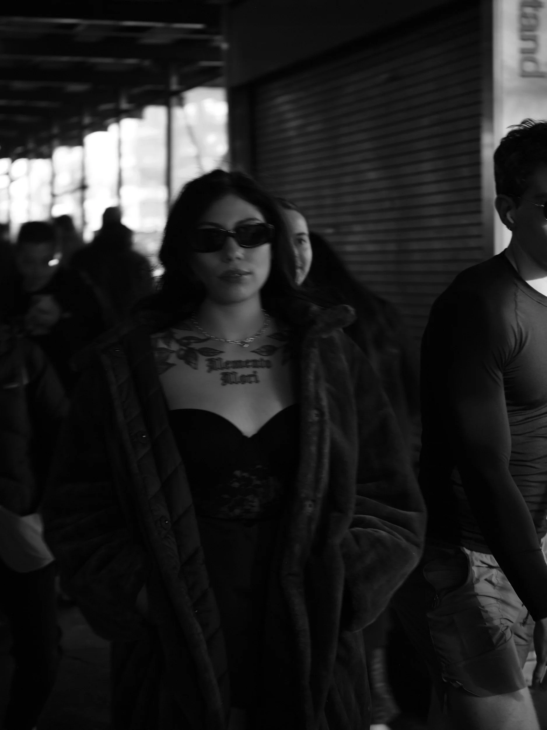 A woman wearing dark sunglasses, a necklace, a fitted black top, and a fur coat walking among a crowd in an urban area.
