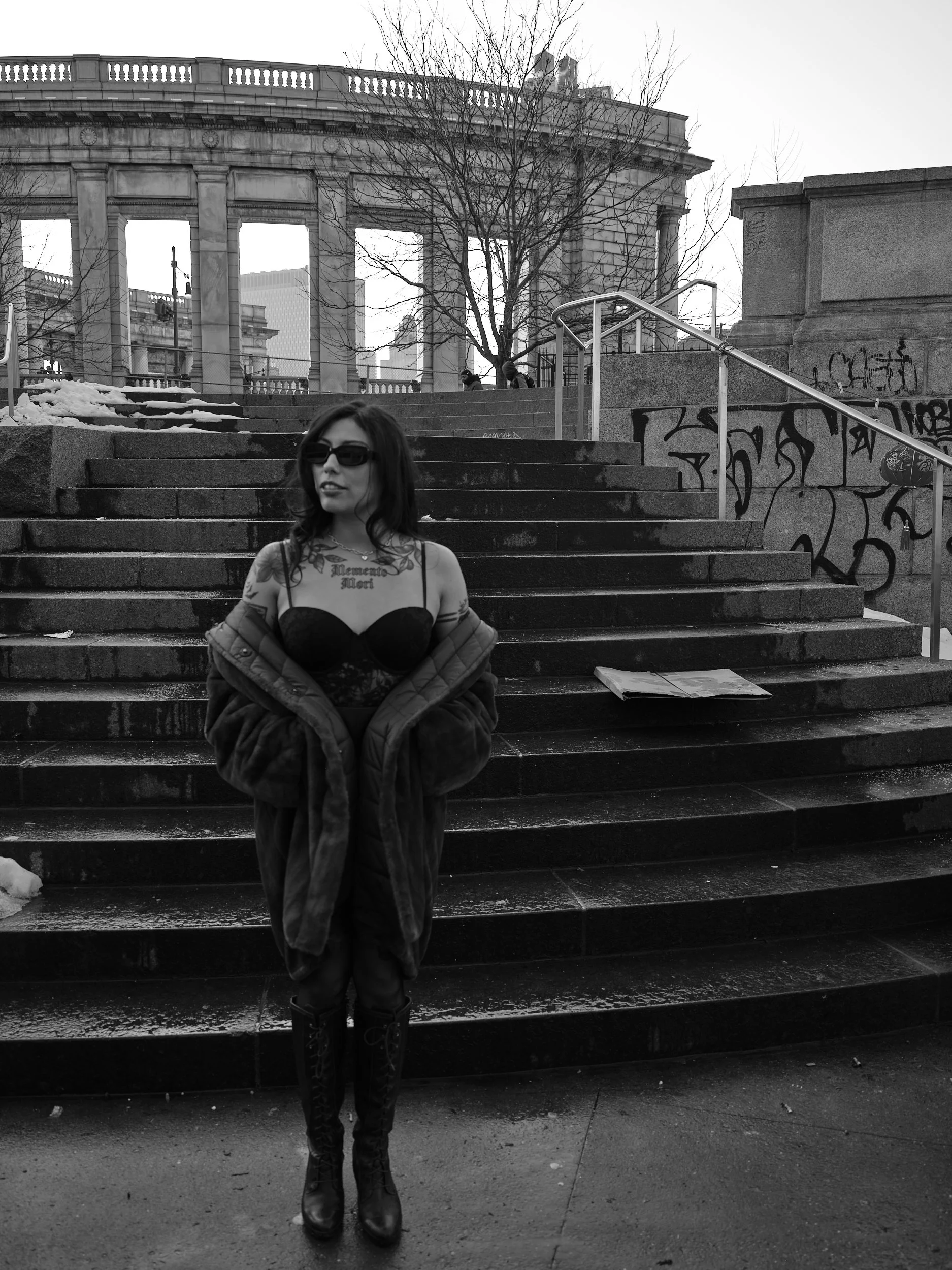 A woman with dark hair, sunglasses, wearing a black corset, lace top, and knee-high black boots, stands on outdoor stone stairs in front of an urban cityscape with a classical building, railings, graffiti, and leafless trees in black and white.