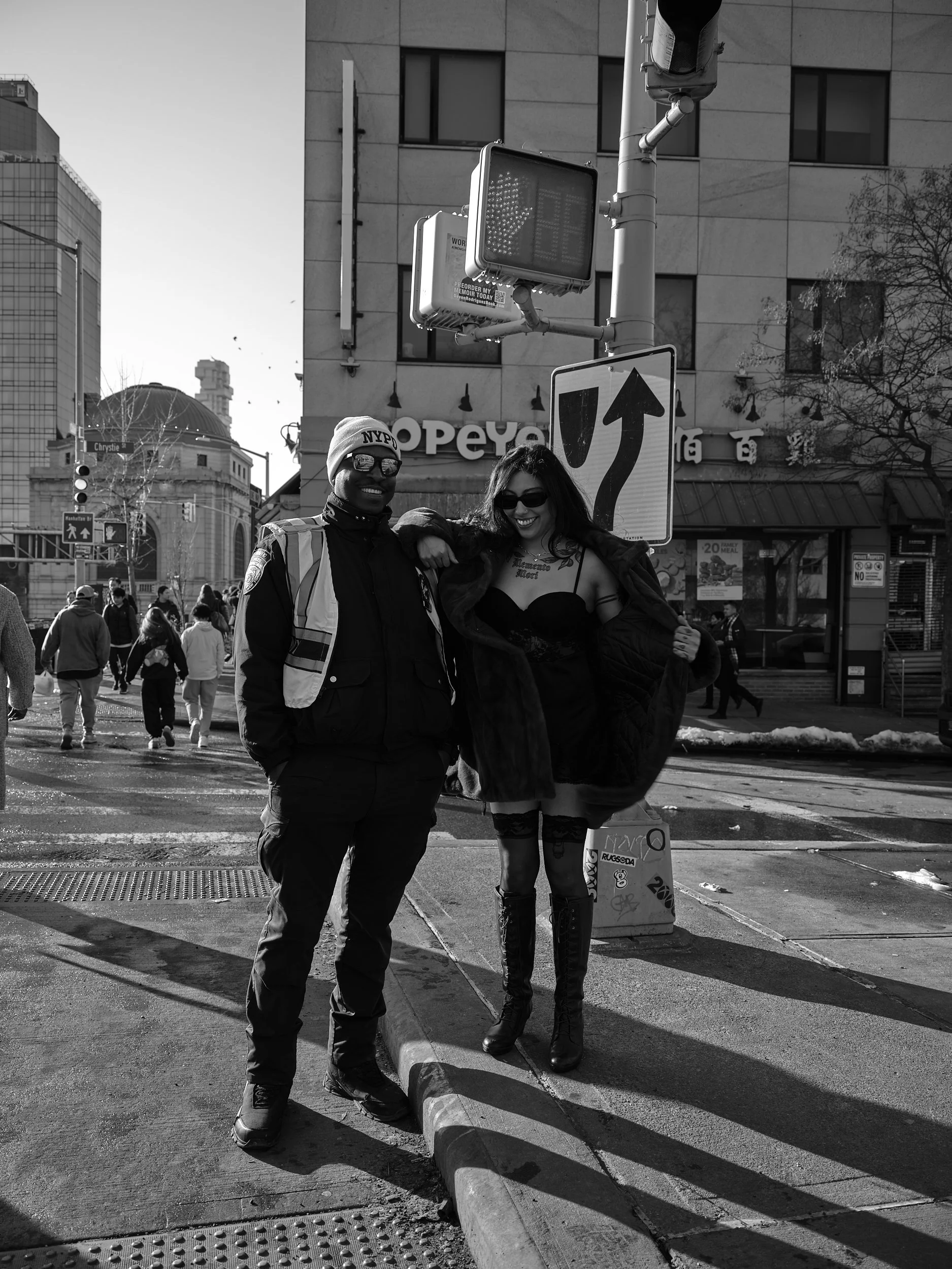 Two people posing on a city street corner, one in a police uniform and the other in a revealing outfit, with buildings and pedestrians in the background.
