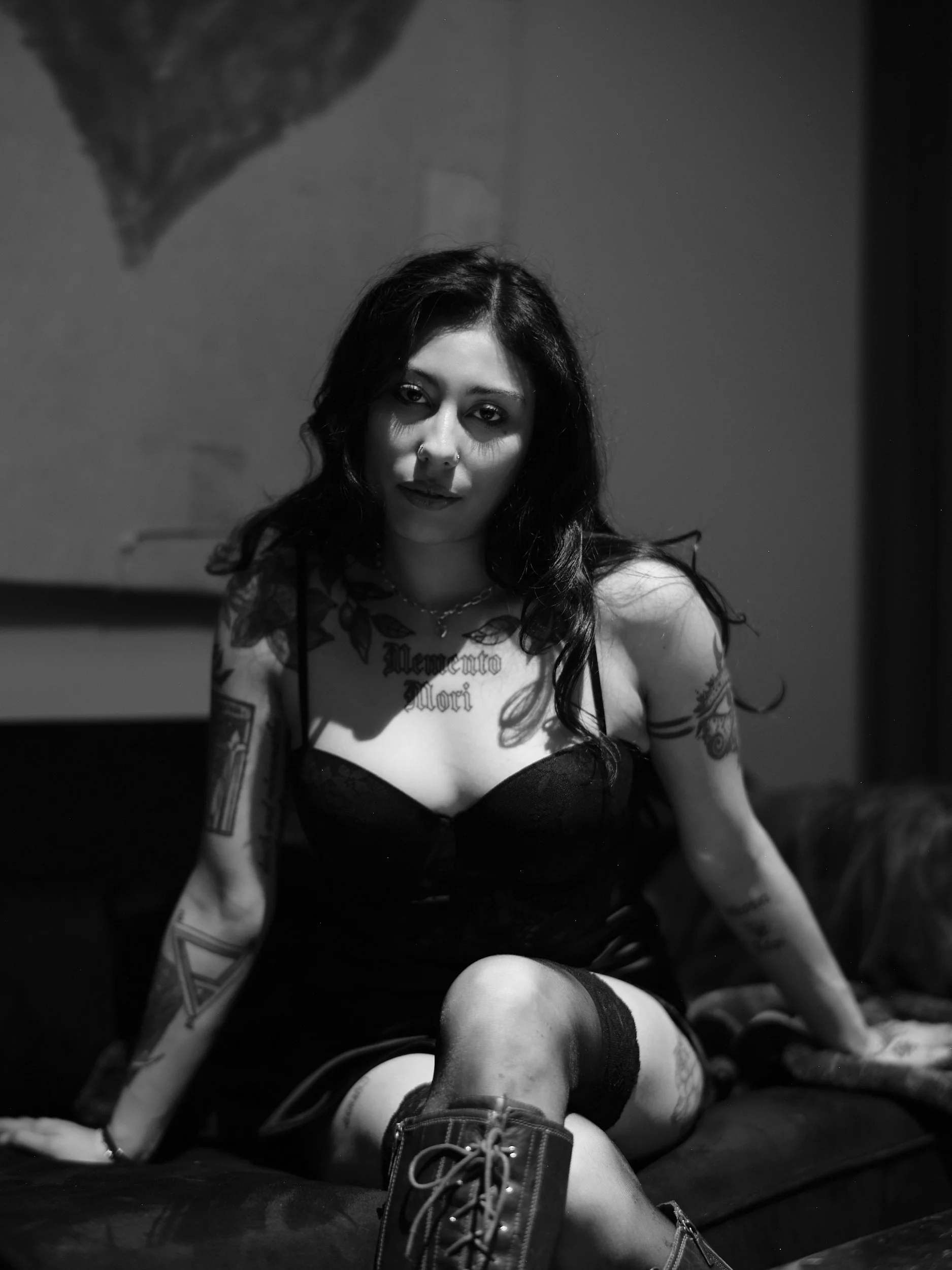 Black and white photo of a woman with dark hair, tattoos, wearing a black dress, sitting on a bed with one knee up, with a serious expression in a dimly lit room.
