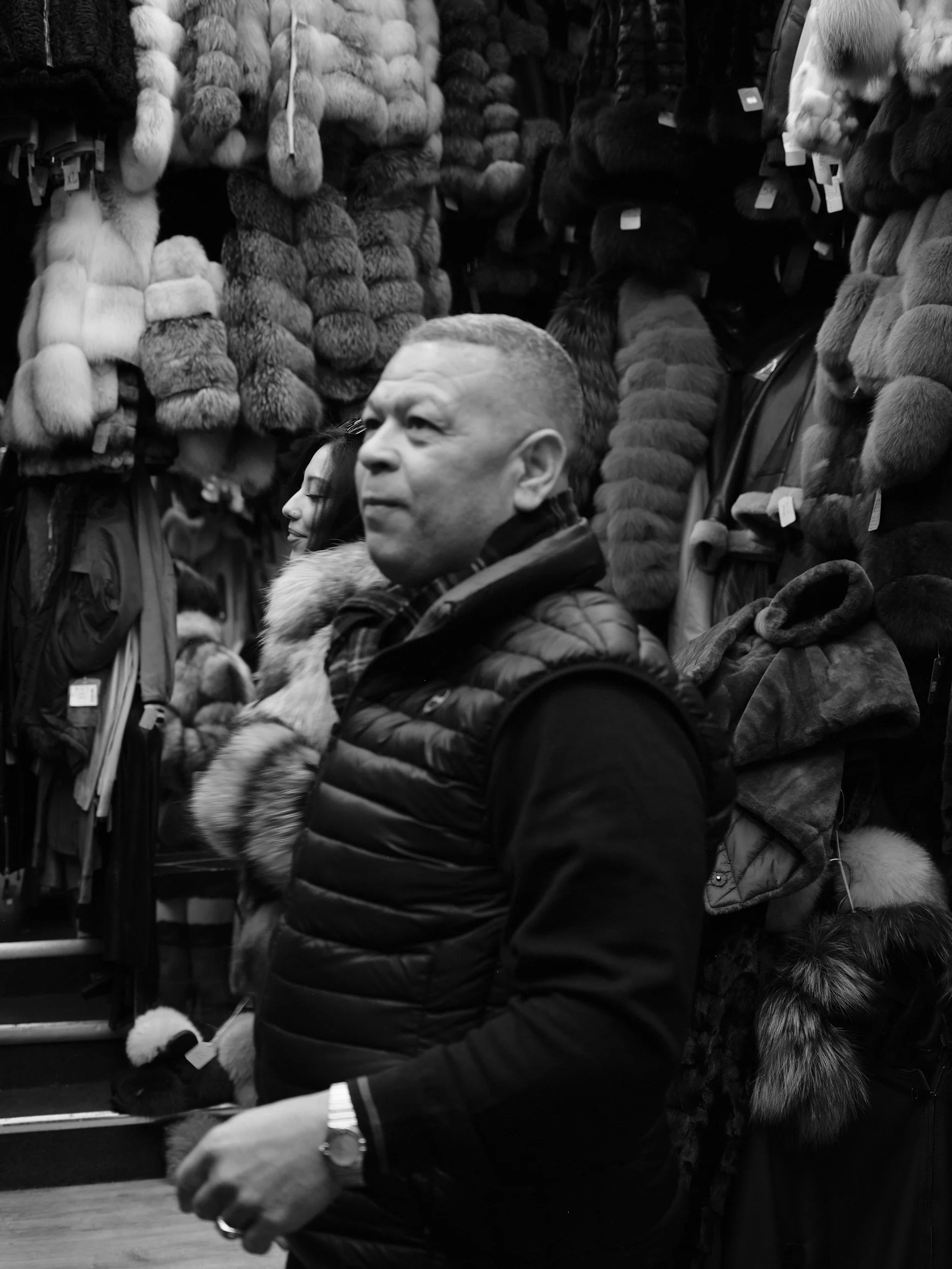 A man in a quilted jacket shopping for fur coats at a store, with fur hats and coats hanging on the wall behind him.