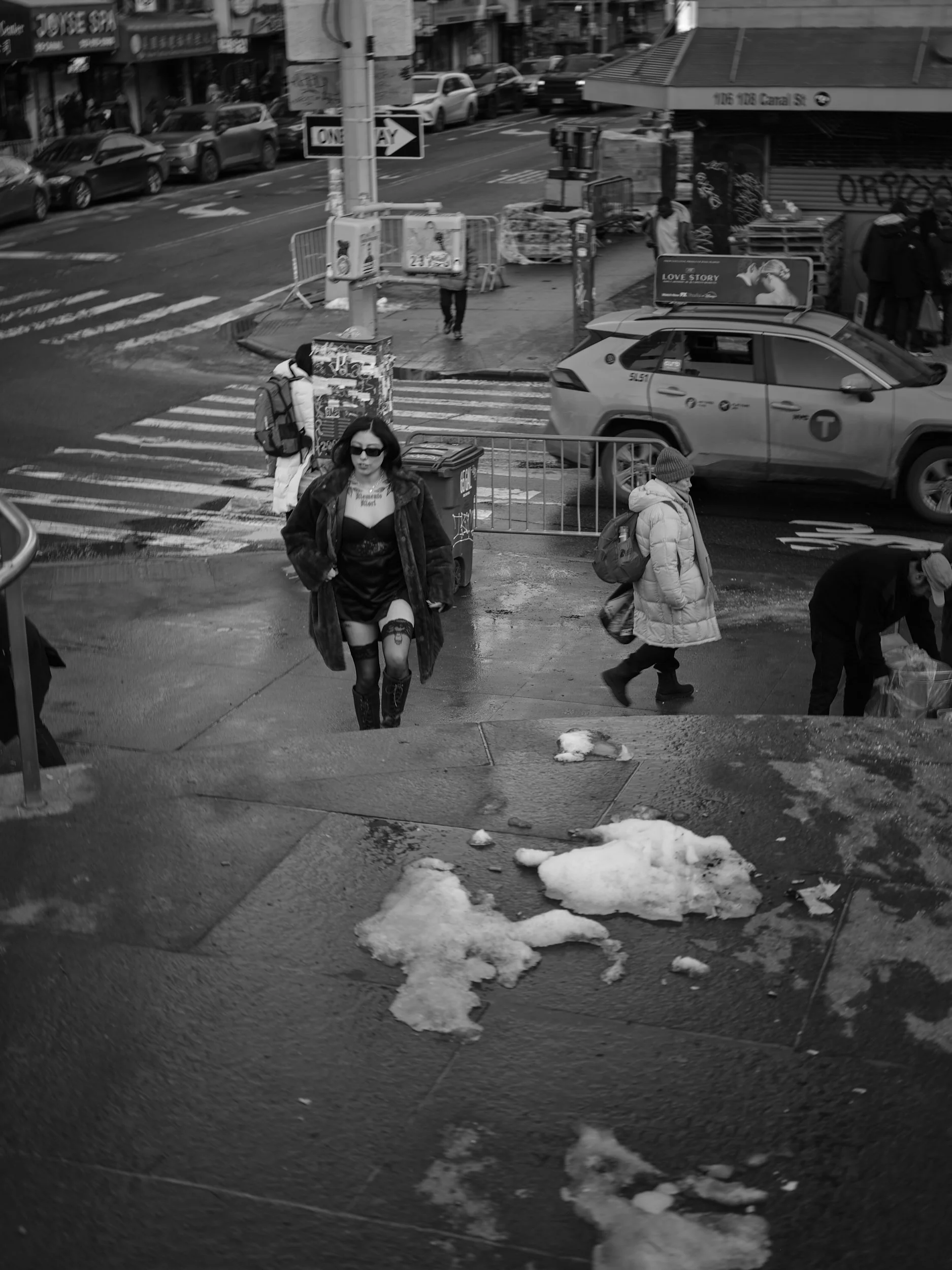 Black and white photo of a city street scene with pedestrians, cars, and buildings. A woman in a coat and sunglasses is walking up a wet sidewalk with patches of snow.