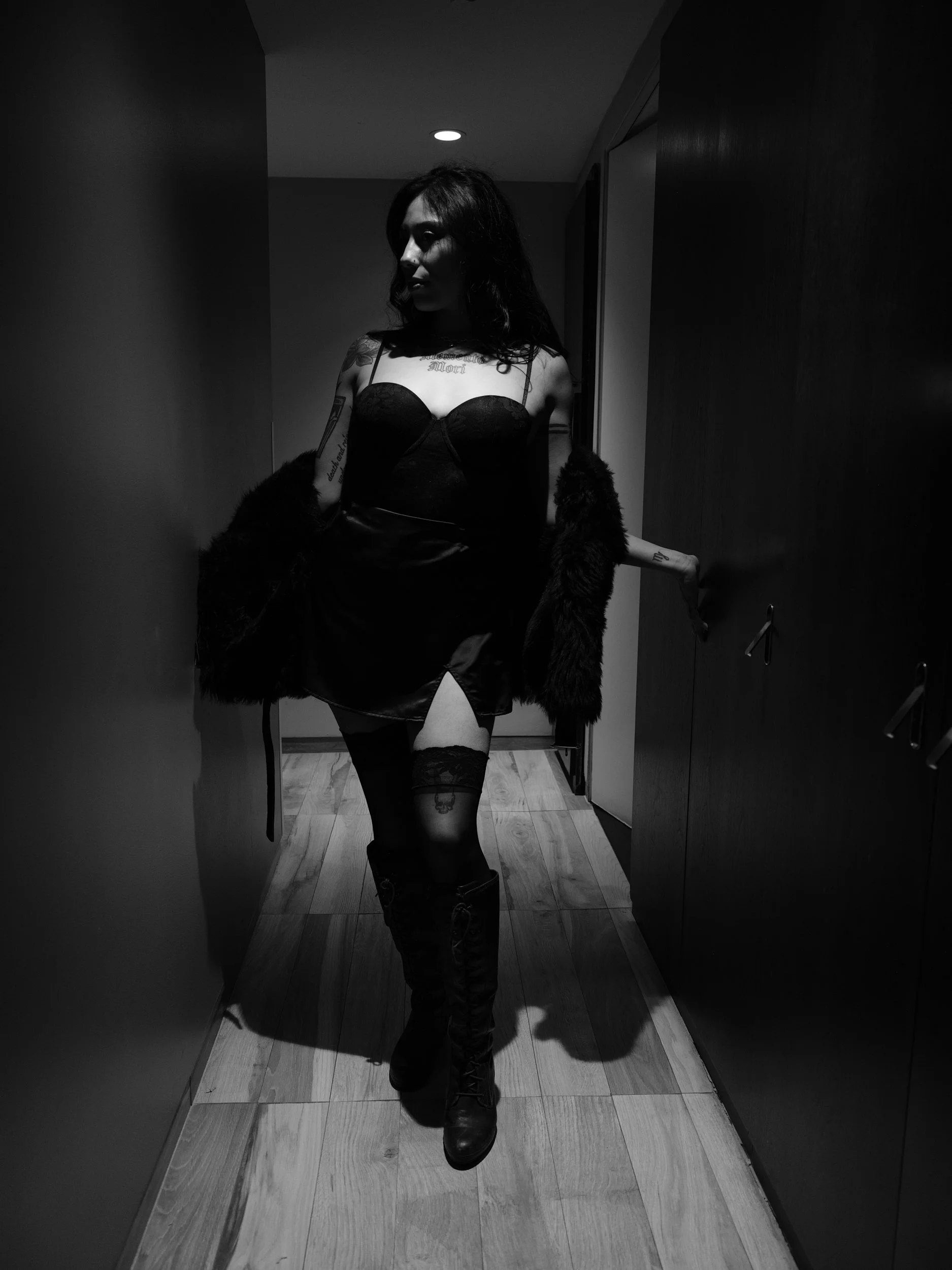 A woman in black lingerie, thigh-high stockings, and boots, standing in a dimly lit hallway and holding a fur coat.
